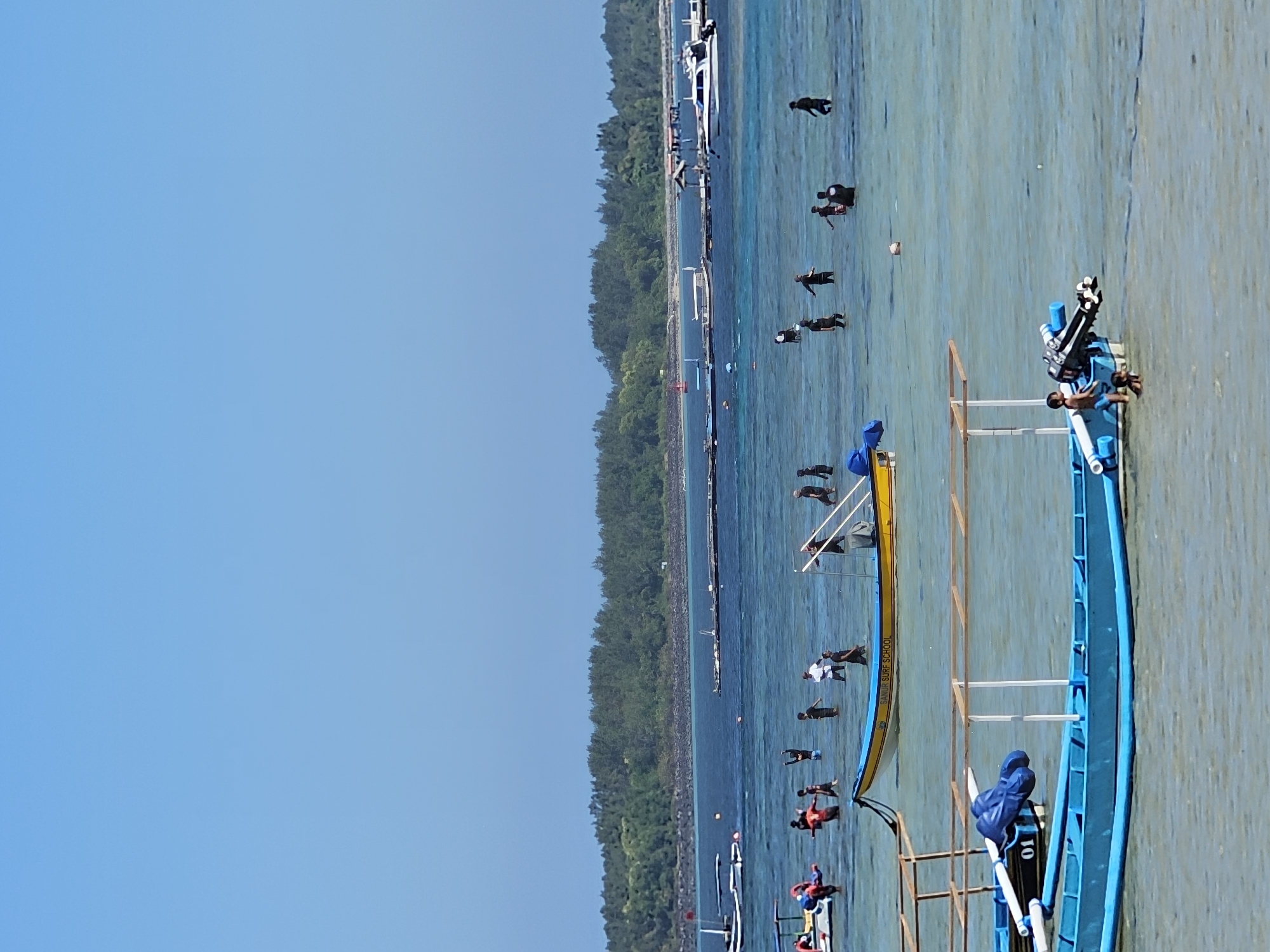 This image captures a lively scene at a beach in Denpasar, Indonesia. The water is clear and shallow, with a sandy bottom visible in some areas. Several boats are present, including a bright blue one with an outboard motor in the foreground, and a yellow and white boat with "SANUR SURF SCHOOL" written on its side further back. A group of people, likely a mix of locals and tourists, are enjoying the water. Some are wading, others appear to be playing or engaging in water activities. In the background, a lush green coastline with dense vegetation stretches across the horizon, bordered by a clear blue sky. The overall atmosphere is one of recreation and leisure on a sunny day.