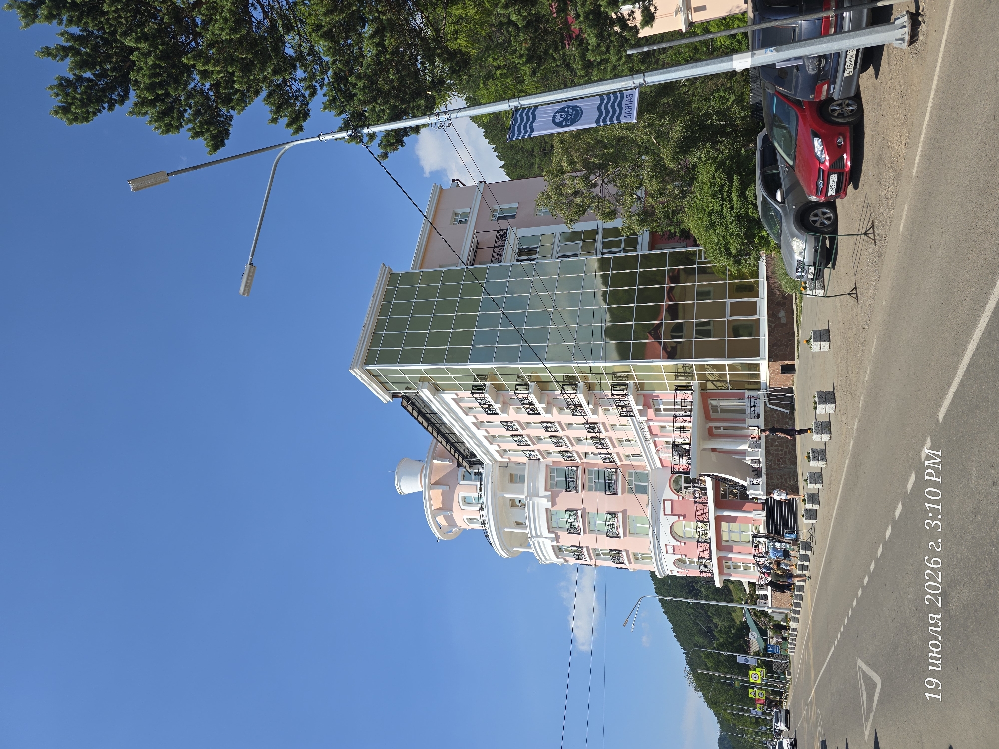 A wide shot captures the pink and white facade of the "Baikal" hotel in Listvyanka, Russia, on a clear, sunny day. The building features a prominent curved section with balconies and a circular turret. Adjacent to it, a modern glass-encased section reflects the surroundings. A streetlamp with two lights stands in the foreground to the right, adorned with a blue and white banner displaying the word "BAIKAL" in reverse. Several cars are parked along the road in front of the hotel. A few people are seen walking near the hotel entrance and along the sidewalk. The road is paved with white markings. In the background, a forested hill rises, suggesting the hotel's location near nature. The bottom left corner of the image displays the timestamp "19 июля 2026 г. 3:10 PM".