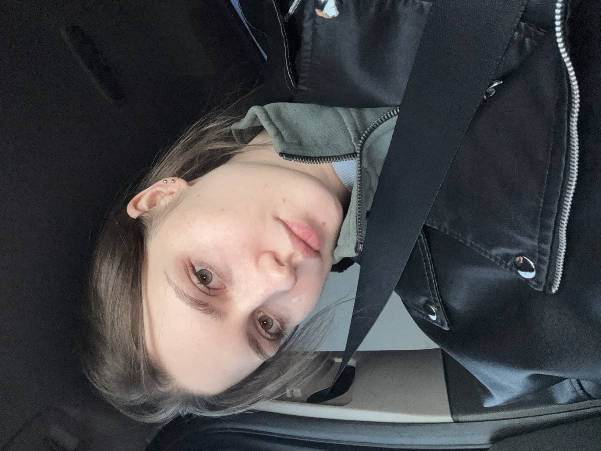 A young woman with fair skin, light brown hair, and brown eyes is wearing a black leather jacket over a gray hoodie and a seatbelt. She is inside a car, with the interior visible. The location is unknown and there is no evidence that the image was taken in Russia.