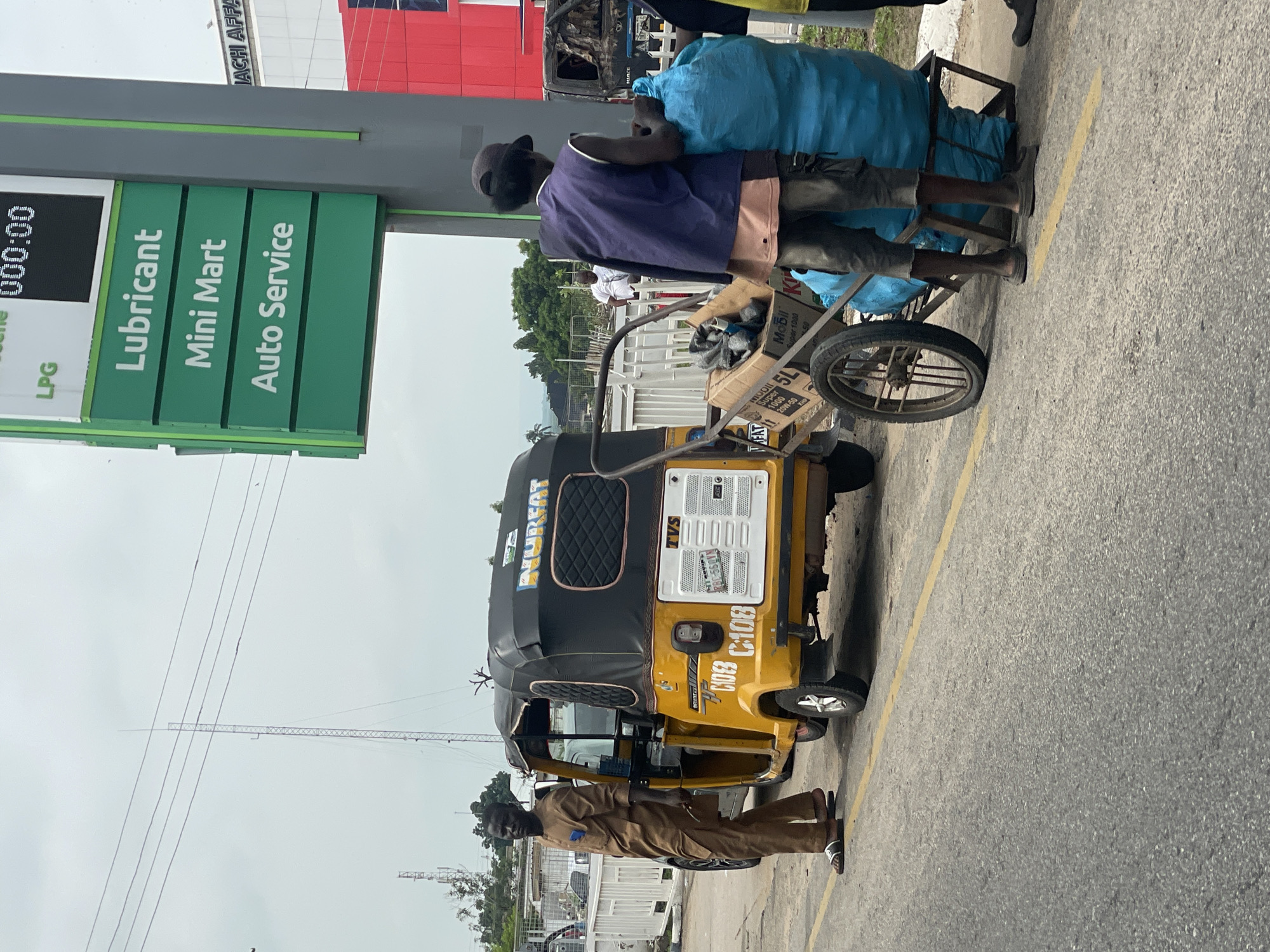 The image captures a street scene in Lokoja, Nigeria, likely near a gas station indicated by a sign listing "LPG," "Lubricant," "Mini Mart," and "Auto Service." In the foreground, a yellow auto-rickshaw, also known as a Keke Napep, is parked on the side of a road marked with a yellow line. A man wearing a traditional brown tunic and pants stands beside the auto-rickshaw, holding a thin object in his right hand.
To the right of the auto-rickshaw, a cart is loaded with several items, including boxes that appear to contain engine oil (indicated by "Mobil 20W-50"). A man wearing a dark vest over a light pink shirt and dark pants is actively engaged with a large, bright blue plastic bag that is part of the cart's load, suggesting he may be loading or unloading it. Another person, partially visible and wearing dark clothing, stands next to him. In the background, more vehicles and structures can be observed, including a tall transmission tower and other buildings, contributing to the urban environment of Lokoja. The sky is overcast.