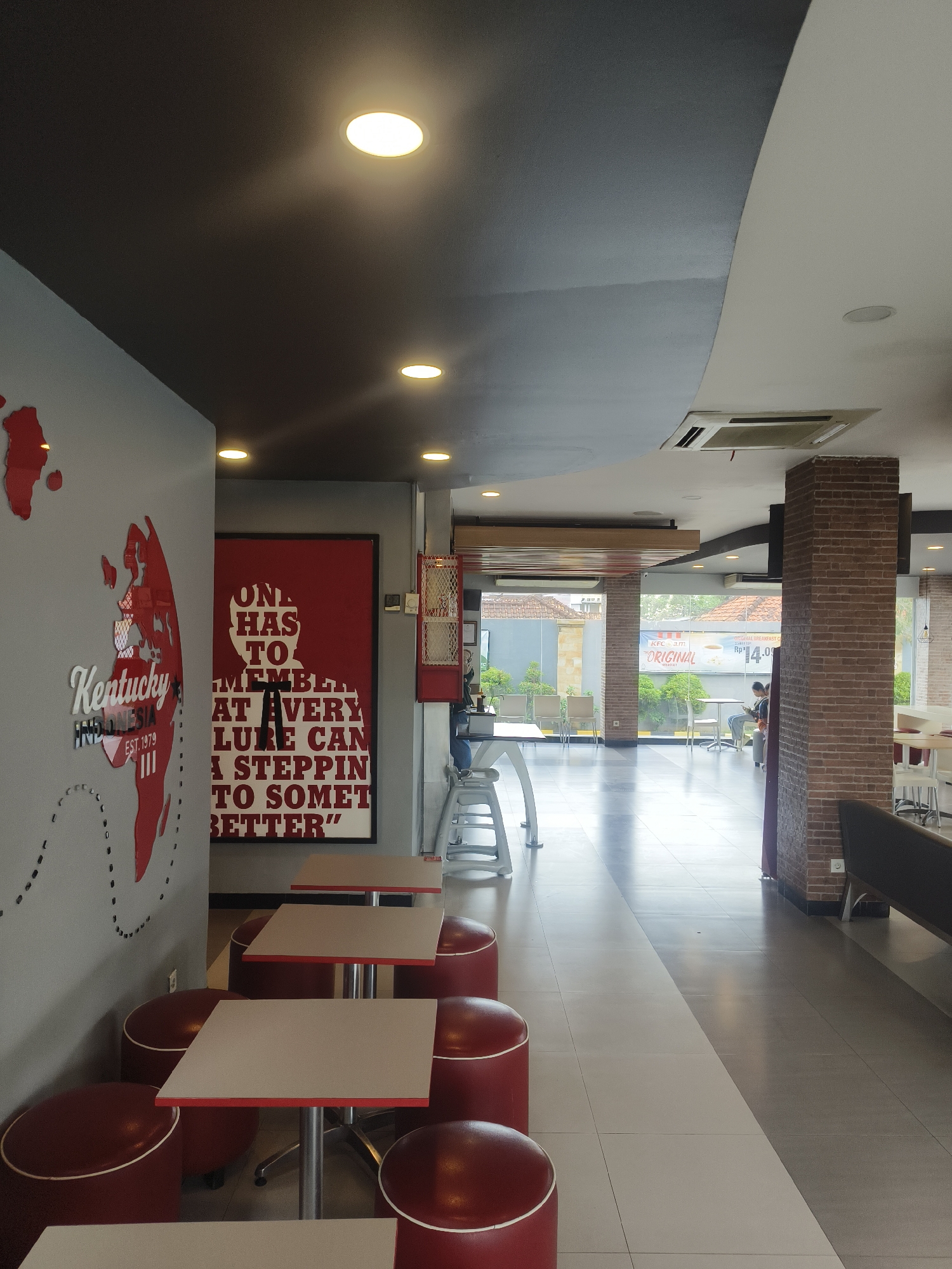 The image shows the interior of a KFC restaurant in Denpasar, Indonesia. The dining area features several tables with red stools. A large red poster with white text is visible on the wall, featuring a quote in English. A map of the world with red outlines is also on the wall, with the word "Kentucky" in stylized white text and "Indonesia EST.1979" below it. Through the glass windows, other diners can be seen seated at tables. The lighting in the restaurant is provided by recessed ceiling lights. The floor is tiled with a contrasting pattern of light and dark tiles. The overall atmosphere is casual and typical of a fast-food establishment.