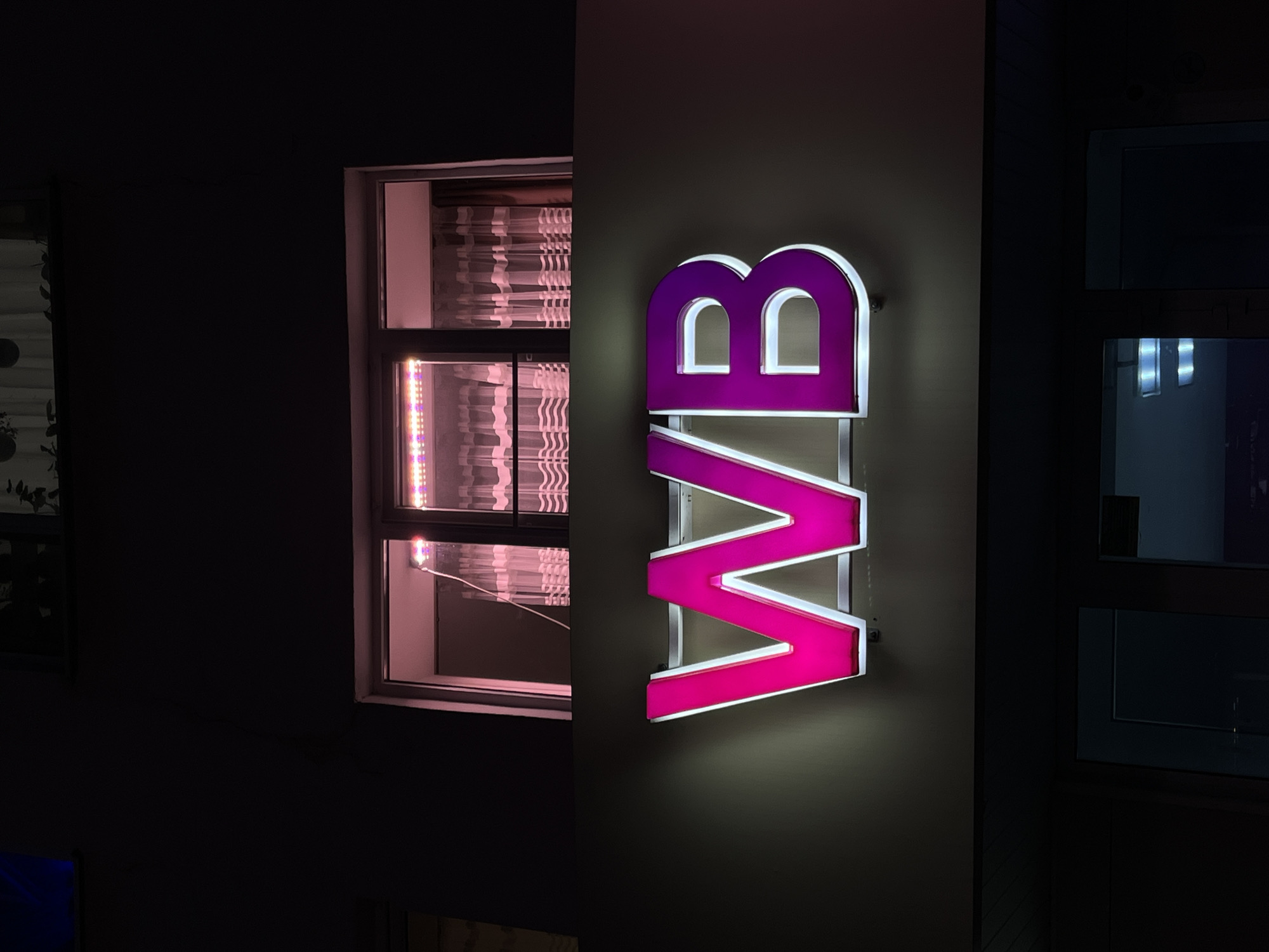 A nighttime shot shows a building exterior in Neftekamsk, Russia, with a brightly lit, vertical sign reading "BWB" in neon pink and purple. The sign is mounted on a wall. To the left is a window with a pink glow coming from inside, appearing to illuminate closed blinds. To the right is another window, dark and reflecting some internal light. The surroundings are dark, suggesting a low-light environment.