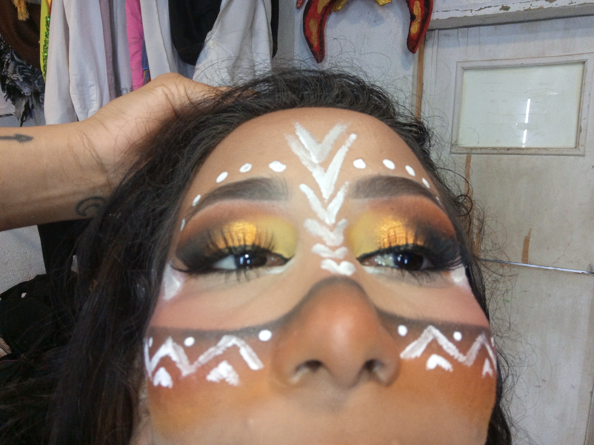 The image is a close-up shot of a person's face with face paint. The paint consists of white geometric designs painted on a tan-colored base. The designs include a series of arrows and dots on the forehead, and zig-zag patterns on the cheeks. The person's eyes are heavily made up with dark eyeshadow and gold glitter. A hand is visible on the upper left side of the image, touching the person's hair. The background includes clothing hanging behind the person. The location is described as Porlamar, Venezuela. The image is of high resolution.