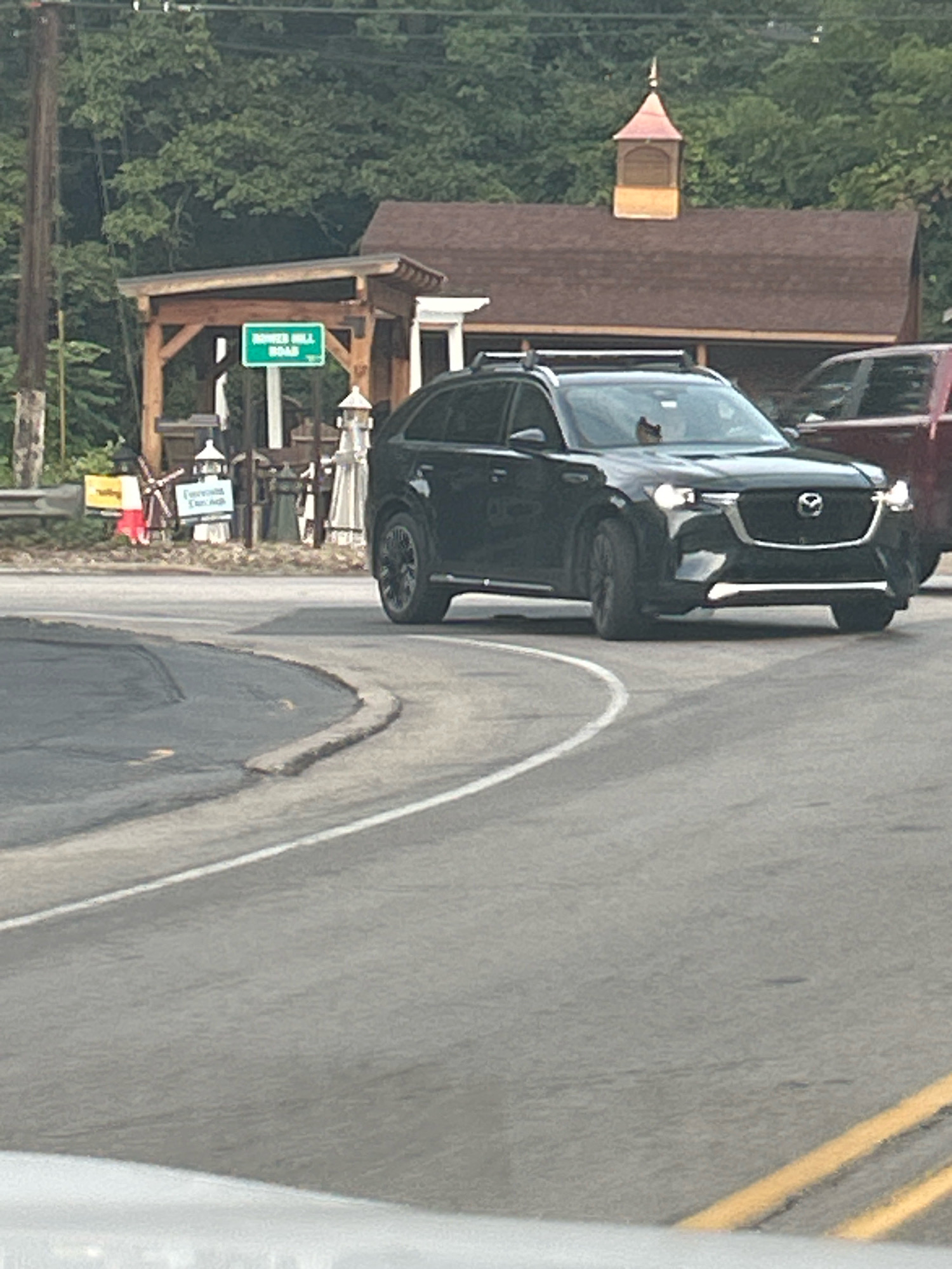 A black Mazda CX-5 SUV is driving on a road that curves to the left. The SUV has its headlights on and roof rails. A dark red SUV is partially visible to the right of the black SUV. In the background, there is a wooden structure with a peaked roof and a cupola. A green sign that reads "BOWERS HILL ROAD" is mounted on a post in front of the wooden structure. Trees and foliage are visible behind the structure. The road surface is asphalt with white and yellow lane markings. The image appears to be taken from a vehicle moving on the same road. The scene suggests an outdoor location, possibly a rural or suburban area.
