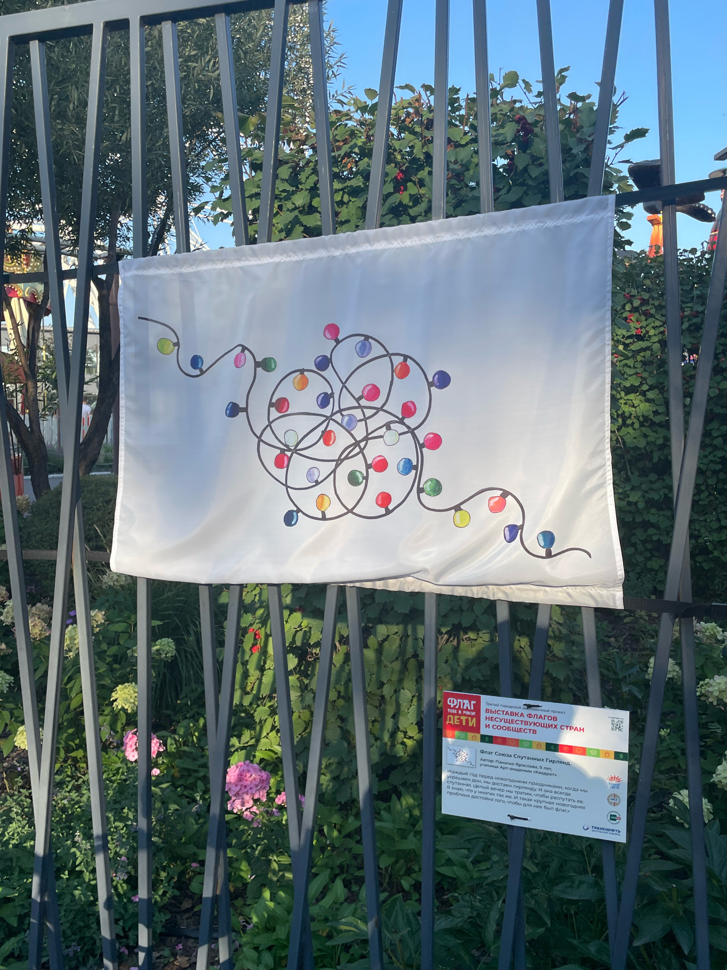 A white flag depicting a tangled string of colorful lights hangs on a dark metal fence. The flag is positioned slightly off-center to the right of the frame. Below the flag, to the right, is a rectangular sign with Cyrillic text and several small colorful icons. Behind the fence, lush green foliage with some pink flowers is visible, suggesting an outdoor setting with vegetation. The lighting suggests it is daytime, with sunlight casting shadows. The sign is a description of the flag and its creator as part of an exhibition. The exhibition title translates to "Flag Exhibition of Non-Existent Countries and Communities". The flag itself is titled "Flag of a String of Christmas Lights" and was created by a 9-year-old student named Panasko Yaroslava. The text on the sign describes the flag as representing the joy and tradition of decorating for holidays, and how it brings people together. The location of this exhibition is the city of Omsk, Russia, specifically within the "Art-Residence" project.