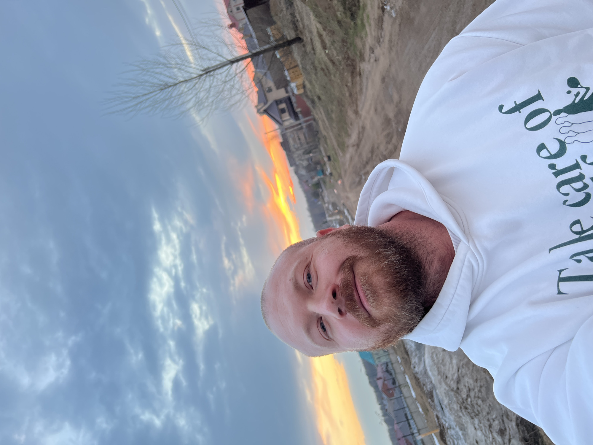 A man with a beard and wearing a white hoodie is posing for a selfie outdoors at Noviy, Russia. The sky is filled with clouds and a streak of orange from the setting sun. A bare tree is visible behind him, along with some houses in the distance. The ground appears to be a mix of dirt and possibly some snow or ice. The text "Take care of..." is visible on his hoodie.