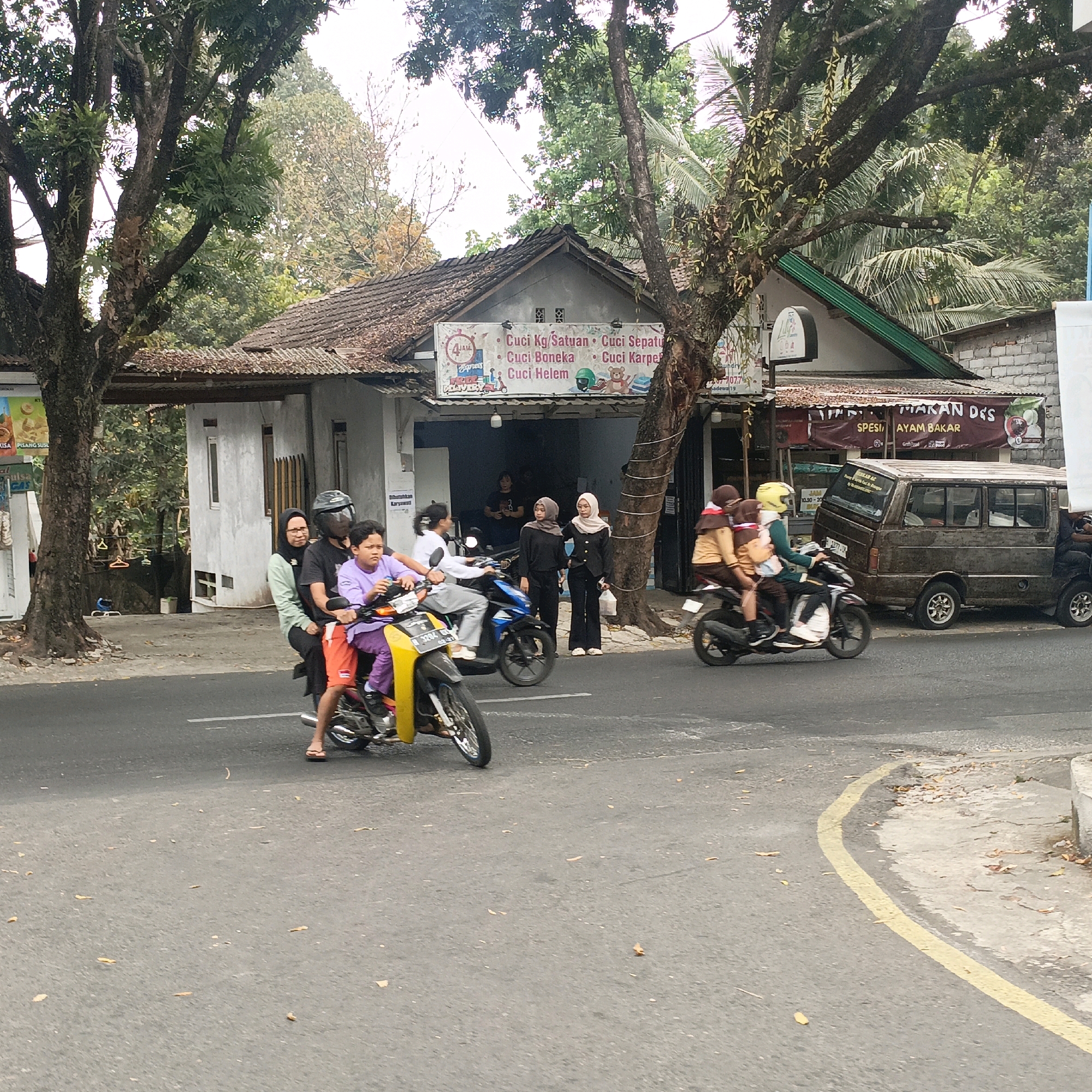 The image depicts a street scene in Salatiga, Indonesia, featuring several individuals on motorcycles and a parked van. In the foreground, a yellow motorcycle carries a woman in a black hijab and a child in a purple shirt and orange shorts. The child is looking to the right. Behind them, another motorcycle is ridden by a person wearing a black hijab and glasses, with a passenger in a light green shirt.
Further down the street, a black motorcycle transports two passengers wearing beige and brown school uniforms, one of whom has a yellow helmet. A dark brown van is parked on the right side of the street.
The background includes buildings with tiled roofs and a banner advertising "MAKAN D'S SPESI AYAM BAKAR" (Eat D's Grilled Chicken Specialties). Trees with lush foliage are visible on both sides of the street, and the sky is overcast. The road is asphalt with a yellow painted line on the right side, and scattered fallen leaves are present on the road and the adjacent curb. The overall impression is of a typical urban street in Indonesia with ongoing traffic and commercial activity.