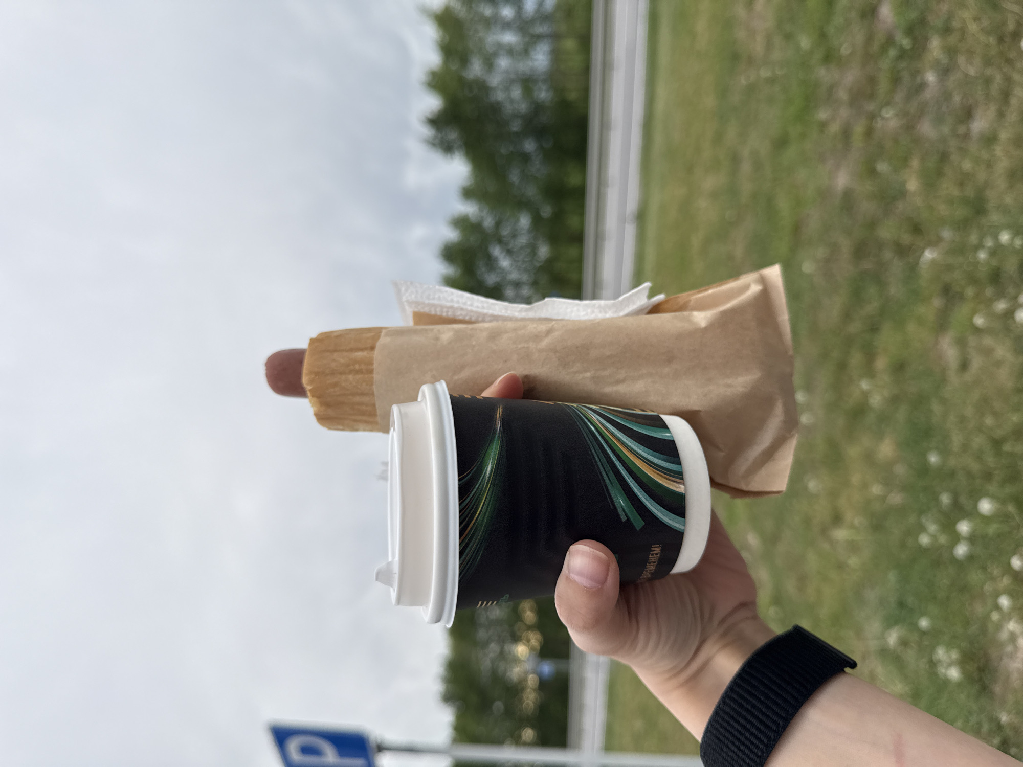 A person holds a cup of coffee and a hot dog in front of a blurred background consisting of grass, trees, and a cloudy sky. The coffee cup is black with a white lid and green and gold accents. The hot dog is wrapped in paper. The person is wearing a black watch. A blue and white parking sign is partially visible in the lower left corner. The Russian word "Паркинг" is printed on the coffee cup. This suggests the location is in Russia. The precise city is unclear, but it is designated as Unknown.