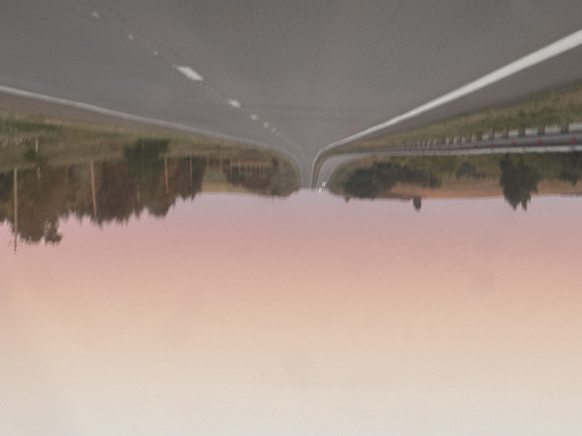 The image depicts a slightly blurry, upward-sloping asphalt road. The sky exhibits a gradient of soft pink and light peach hues, suggesting either dawn or dusk. Trees and fields border the road. The road is marked with a solid white line on the left and dashed white lines on the right. In the distance, at the crest of the hill, two faint white lights, possibly from headlights of an oncoming vehicle, are visible. The scene is devoid of any people or discernible human activity beyond the presence of the road and the distant vehicle lights. The environment appears to be rural or semi-rural. The location is identified as Pridorozhny, Russia.