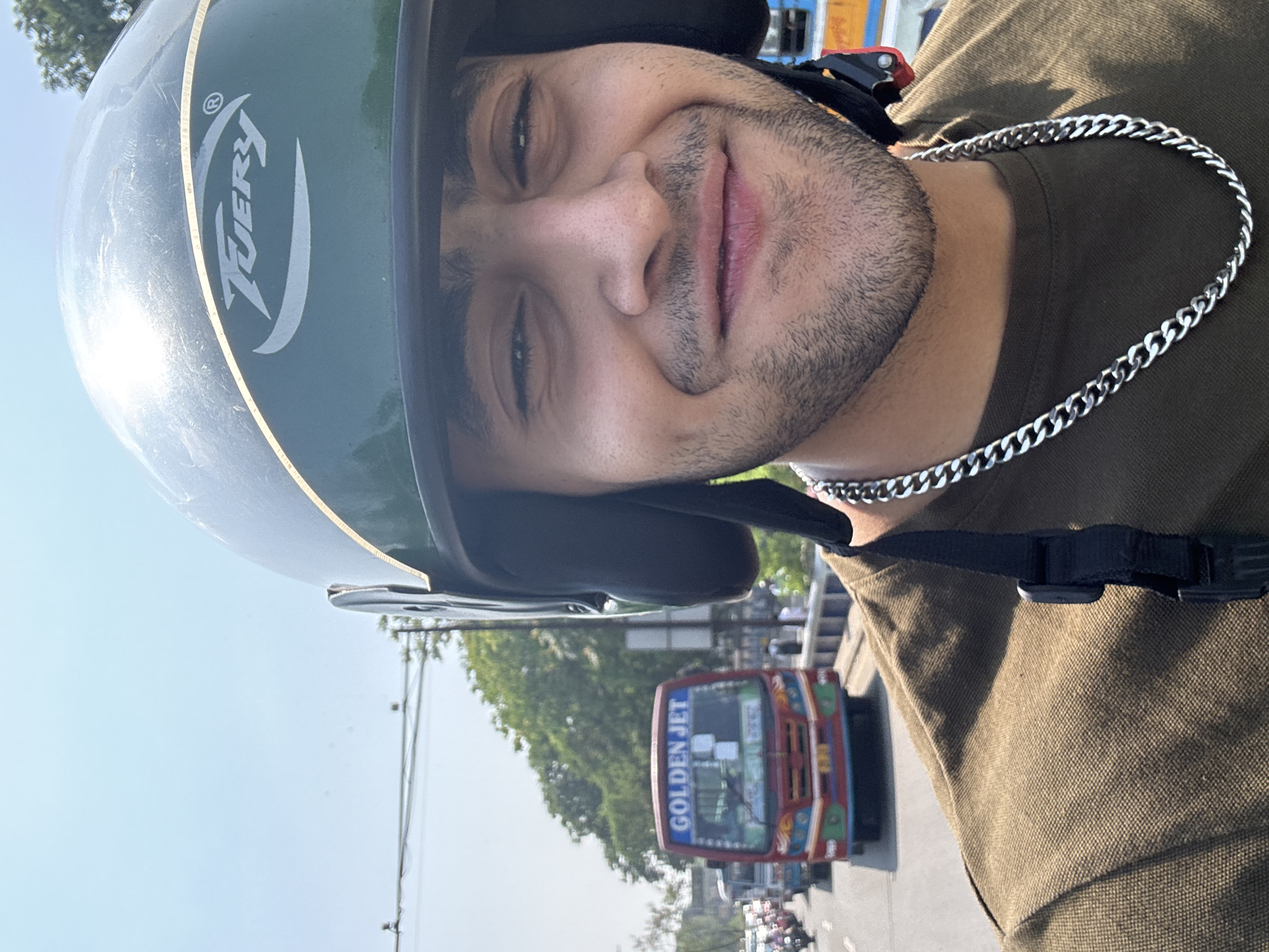 A close-up, eye-level, selfie-style photograph depicts a young adult male wearing a dark green "FUERY" brand motorcycle helmet with a golden stripe and an olive green polo shirt, accented by a silver curb-link chain necklace. He has short dark hair visible from the sides, stubble on his face, and a slight smile directed towards the camera.
The background shows a busy urban street scene in daylight. A prominent red public transport bus with "GOLDEN JET" in white capital letters on its destination board, featuring blue, green, and yellow trim, is positioned in the mid-ground. Numerous other vehicles, including motorcycles and cars, are visible in traffic behind the bus. The road appears to be part of a bridge or elevated structure, evidenced by a concrete railing visible on the right. Green trees and overhead power lines are present against a clear blue sky.
The distinctive "GOLDEN JET" bus and the general urban environment confirm the location as Kolkata, India.