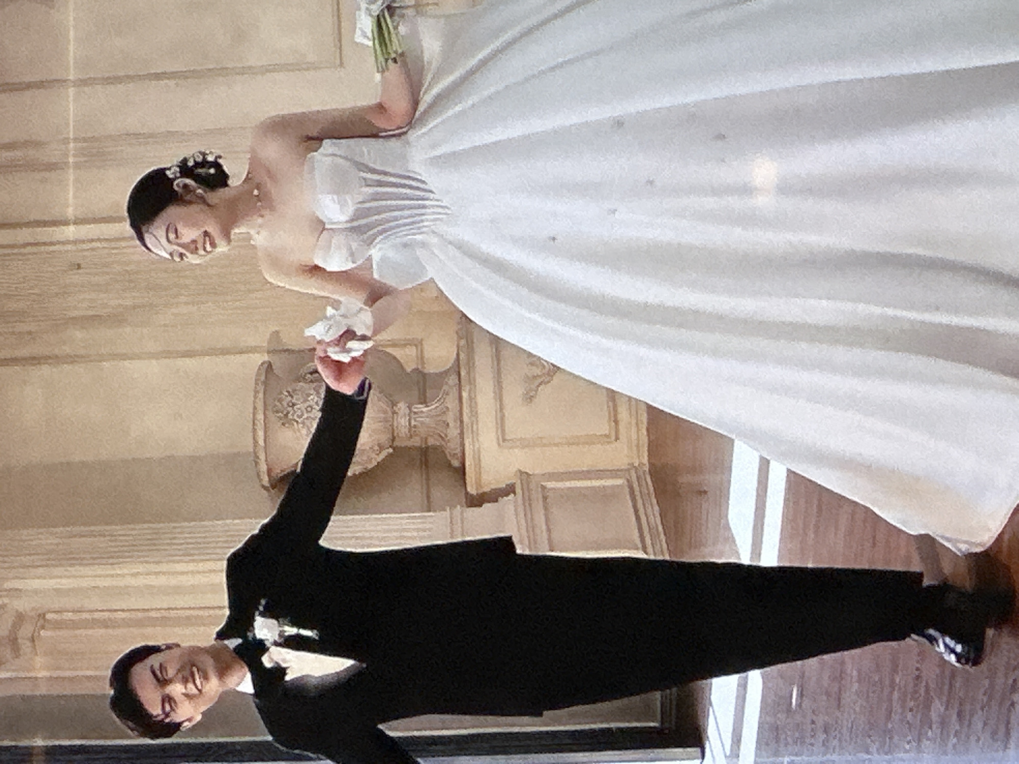 A wedding portrait depicts a smiling couple. The bride, on the left, is wearing a strapless white wedding gown with a full skirt, a delicate necklace, and flowers in her hair. She is holding a bouquet. The groom, on the right, is dressed in a black suit with a white shirt and a bow tie. He is extending his left hand toward the bride, and they appear to be holding hands. The background consists of a room with light-colored walls and architectural details. The photograph is taken indoors, possibly in a grand hall. This is likely not located in Bac Nha Trang Ward, Vietnam.
