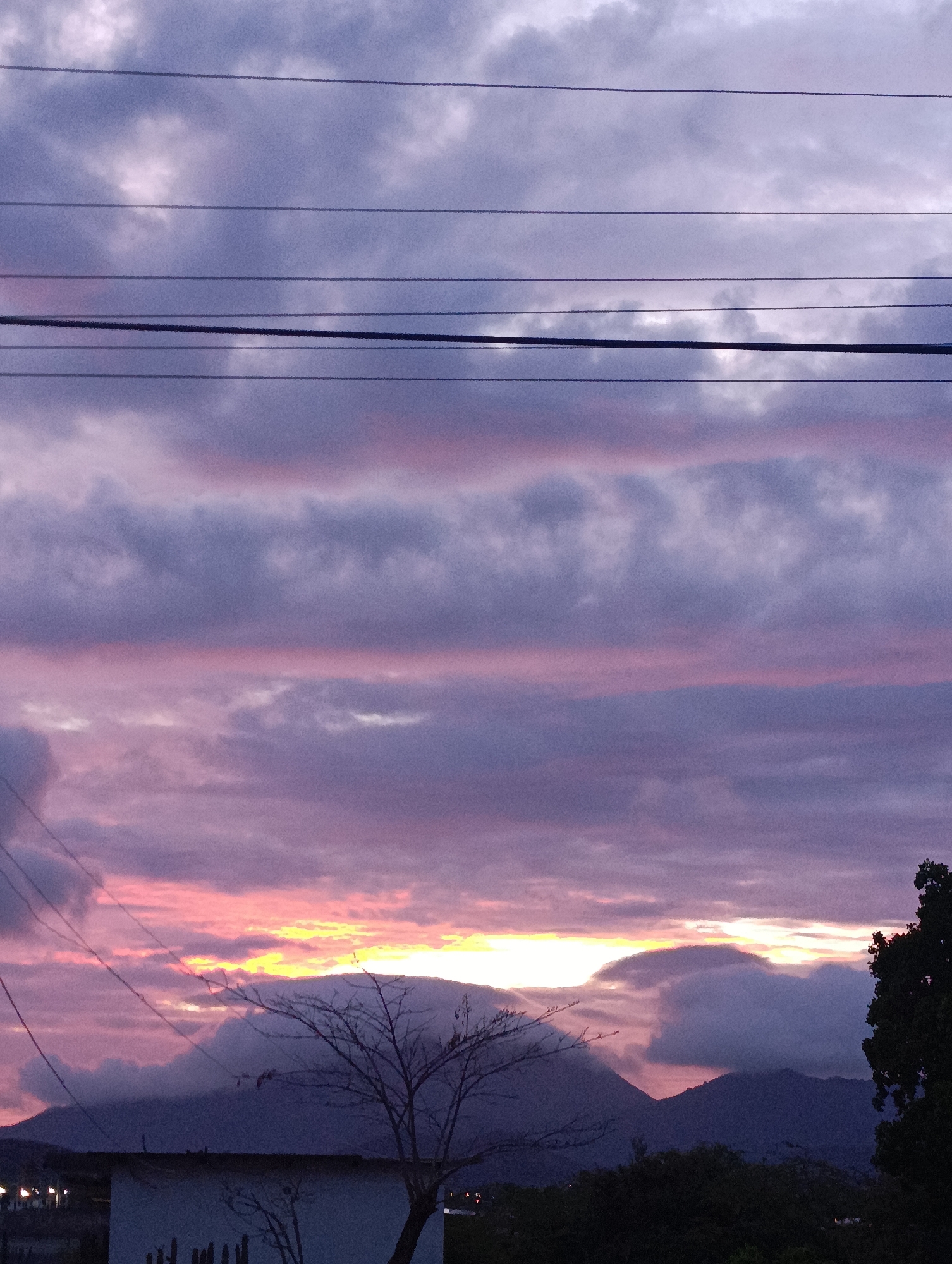 The image depicts a sunset in Juan Griego, Venezuela. The sky is filled with layers of clouds, ranging in color from dark grey to pink and purple, with a bright yellow horizon. In the foreground, electrical wires are visible across the top portion of the frame. Below them, there's a bare tree with a mountain range behind it. A rooftop and some foliage are also visible in the lower portion of the frame. Lights suggest there are structures with street lights and/or residence lights and cars on the distant bottom left side.