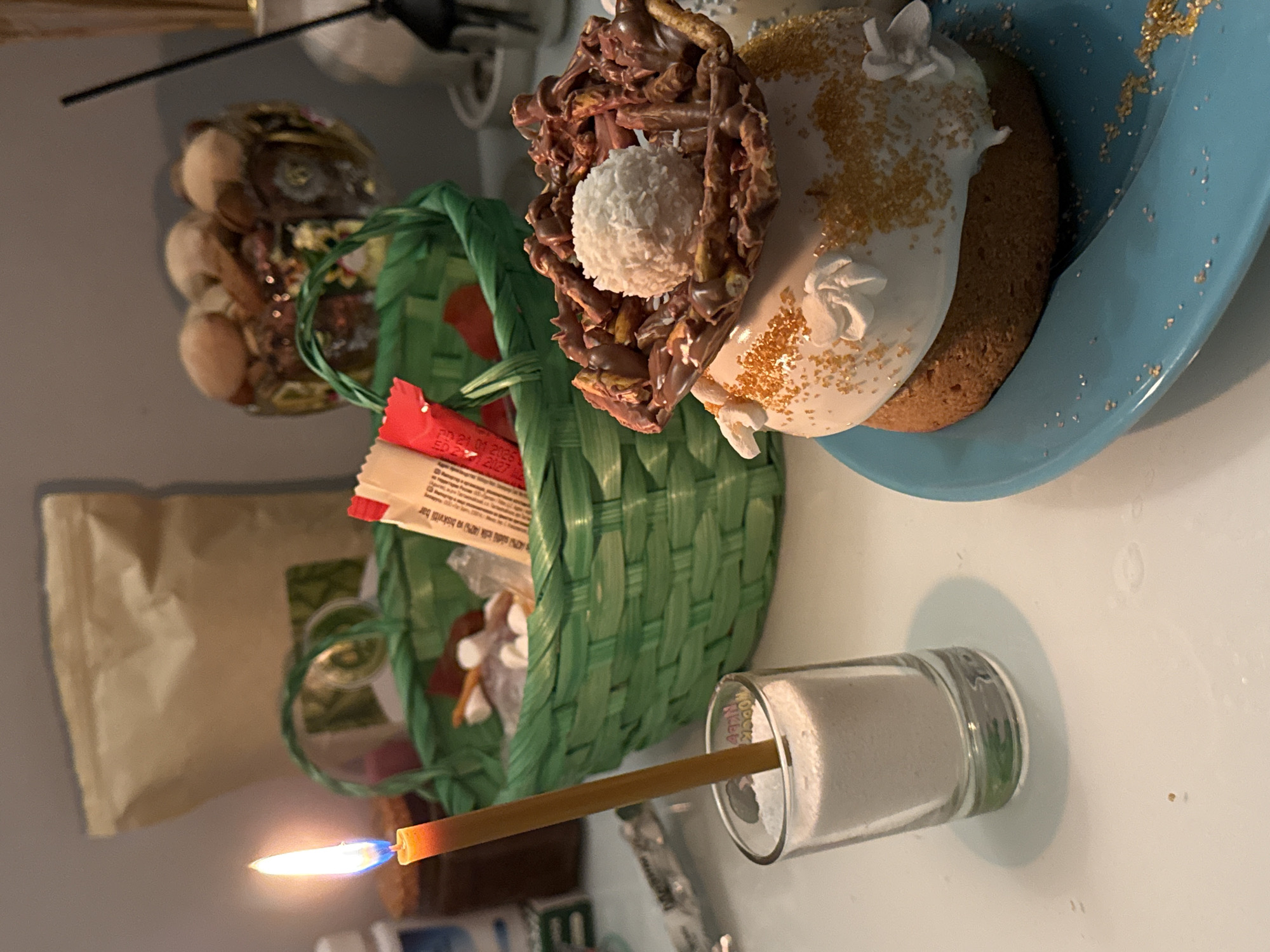 A still-life photograph depicts a partially-filled green woven basket containing items including candies and packaged food. The basket is situated next to a lit beeswax candle inside a clear glass container filled with white material, likely salt or sugar. In the background, there is a partially visible beige paper bag. To the right of the basket, on a light blue plate, sits a decorated cake or sweet bread with white frosting and edible decorations. A glass jar containing miscellaneous items is also partially visible in the background. The photograph appears to be taken in an indoor setting on a light-colored surface. The city and state of Unknown, Unknown cannot be determined from the given information.