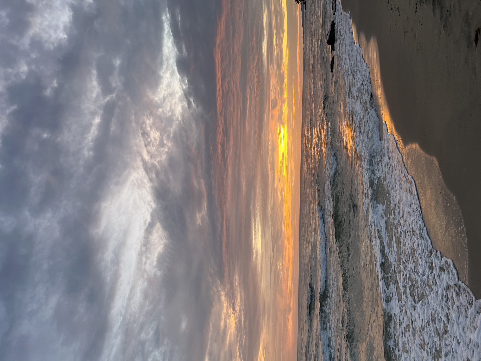 This image captures a serene beach scene at sunset on Phú Quốc, Vietnam. The sky is filled with dramatic, layered clouds painted in shades of gray, pink, and orange, transitioning to a brilliant yellow band of light at the horizon where the sun is setting. The ocean is calm with gentle waves lapping against the shore, their white foam creating a stark contrast with the darker sand. The wet sand reflects the warm hues of the sunset, creating a shimmering path along the waterline. A few dark rocks are visible in the water and on the beach. The overall atmosphere is tranquil and picturesque, showcasing the natural beauty of the coastal landscape.