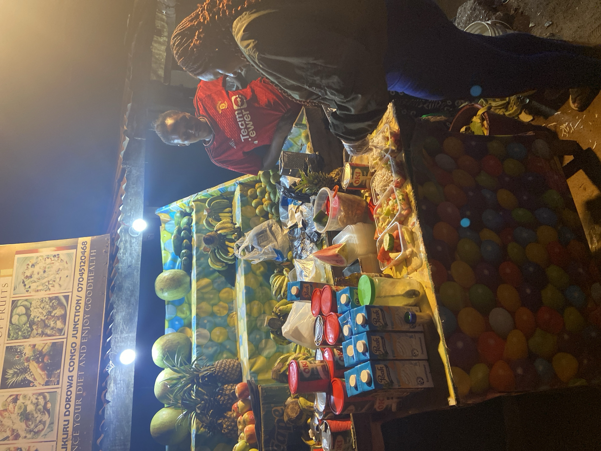 The image depicts an outdoor fruit stand at night, possibly in Du, Nigeria, as suggested by the text on a sign that reads "UKURU DOROWA CONGO JUNCTION / 07045120468". The stand is illuminated by overhead lights. On display are various fruits including pineapples, watermelons, bananas, and what appear to be mangoes or other citrus fruits. There are also packaged juices, canned goods, and pre-packaged fruit salads in clear containers.
Two women are present at the stand. One woman, wearing a red Manchester United jersey with "TeamViewer" printed on it, is behind the counter. Another woman, wearing a dark jacket and blue leggings, is standing in front of the counter, appearing to be served or preparing something. She has her hands in clear plastic gloves and is leaning over the counter towards the fruit salads.
The ground around the stand is dirty, with some debris and discarded materials. The overall scene suggests a bustling street-side vendor at night, serving customers.