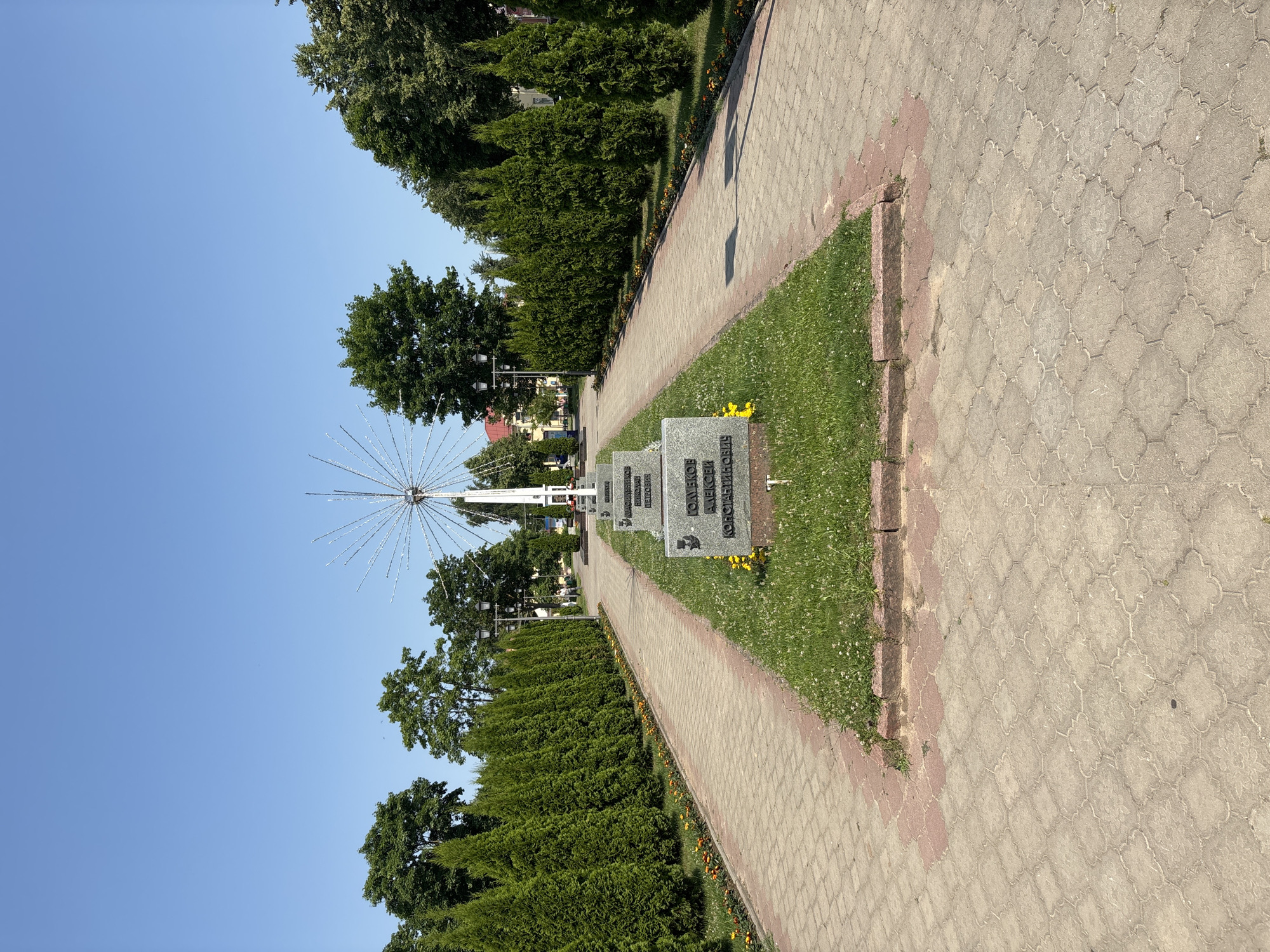 The image shows a memorial in Pastavy, Belarus. The memorial consists of a central pedestal with several granite slabs inscribed with names, leading to a larger monument with a spiky, star-like structure at its apex, possibly illuminated by lights. The memorial is situated in a park-like setting with rows of manicured green bushes flanking a paved walkway. The walkway is made of interlocking pavers in a light tan color with a red border. Small orange flowers line the edges of the walkway. The sky is clear and blue, and lush green trees are visible in the background. A few people are walking in the distance along the path. The scene appears to be a public space, possibly a park or a memorial garden.