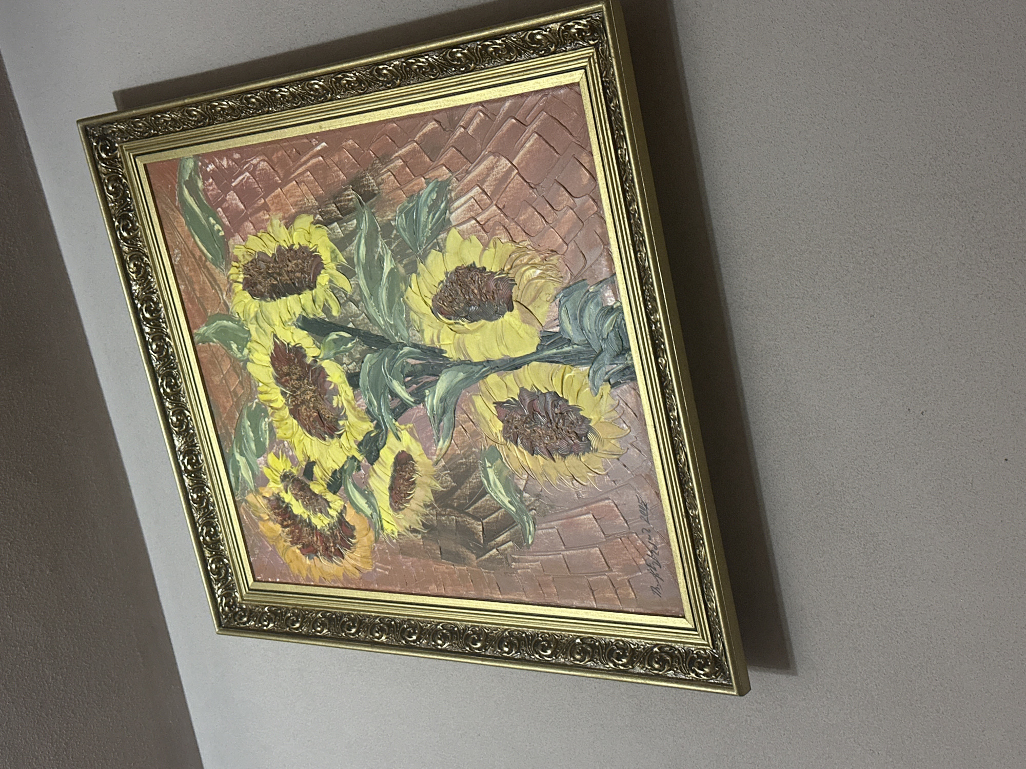 A framed painting of sunflowers hangs on a light-colored wall. The frame is ornate and gold-colored. The painting depicts several sunflowers with yellow petals and dark centers, along with green leaves, against a textured background of brown hues. The image suggests an indoor setting. The location is Unknown, Unknown.