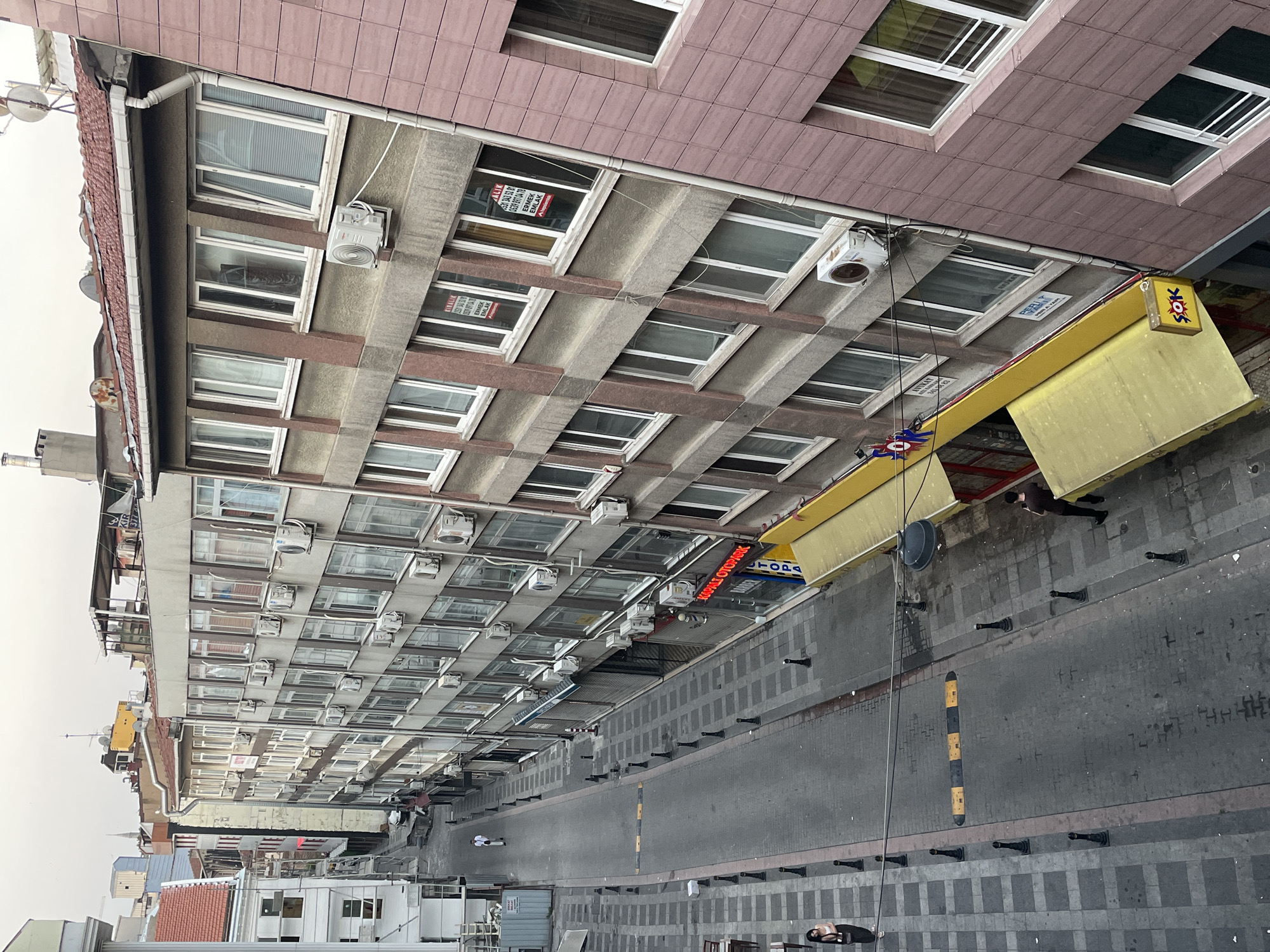 An exterior, high-angle, vertical shot captures a street in Kadıköy, Turkey. The street is lined with multi-story buildings, mostly appearing residential or commercial. On the left side of the street, a sidewalk with a checkerboard pattern of grey tiles leads to a two-story building with white siding and red-tiled roofing. Further down the street, a narrower building with a pale yellow facade and arched windows is visible. The right side of the street features taller, more modern buildings with numerous windows and air conditioning units protruding from the facades. The buildings on the right appear to be a mix of residential apartments and commercial spaces, with some storefronts visible at ground level. A few awnings, one yellow and one red, extend from the buildings. There are several parked cars and a single yellow and black speed bump on the street.
Two women are visible on the sidewalk. One, wearing a white shirt and black pants, stands near the edge of the sidewalk on the left side of the image. The other, in a light-colored top and dark skirt, is further down the street on the left, near a small table and chairs. One man, in a white shirt and dark pants, walks down the street from the upper left of the frame. Another man, in a dark shirt and pants, stands on the sidewalk on the right side of the image, facing away from the camera.
The sky is overcast, suggesting a cloudy day. The overall scene depicts a typical urban streetscape in a Turkish city, with a mix of architectural styles and daily life unfolding.
