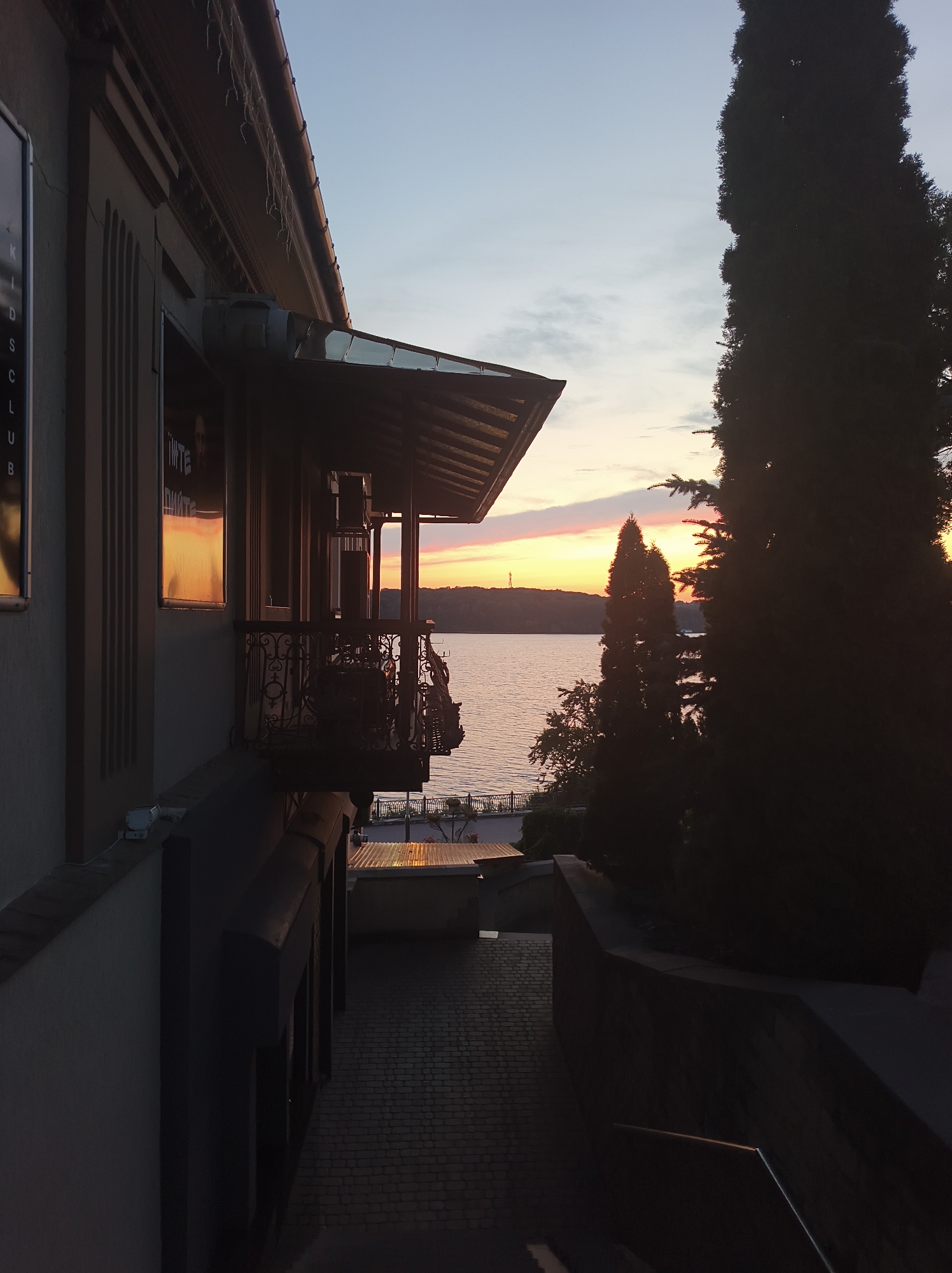 The image depicts a serene twilight scene, likely at dusk, overlooking a body of water, possibly Ternopil Pond, in Ternopil, Ukraine. In the foreground, the left side of the frame is dominated by the facade of a building with a covered balcony. The building's architecture features textured walls and decorative elements, including ornate railings on the balcony. A sign with vertical lettering, partially visible and reading "KIDS CLUB," is affixed to the building. The balcony itself offers a view towards the water and the horizon.
To the right of the building and balcony, a tall, dark, conical tree stands prominently, silhouetted against the sky. Several other trees and foliage are also visible, contributing to the natural setting.
The background features a wide expanse of water, reflecting the soft hues of the setting sun. The sky above is painted with gradients of pale blue and warm orange and pink, indicating the sunset. A distant silhouette of land with some structures, possibly on the opposite shore of the pond, is visible on the horizon, with a tall, thin structure, perhaps a tower or antenna, marking the skyline.
The ground in the immediate foreground, leading away from the building, is paved with interlocking bricks, suggesting a pathway or courtyard. The overall atmosphere is peaceful and tranquil, characteristic of a calm evening by the water.