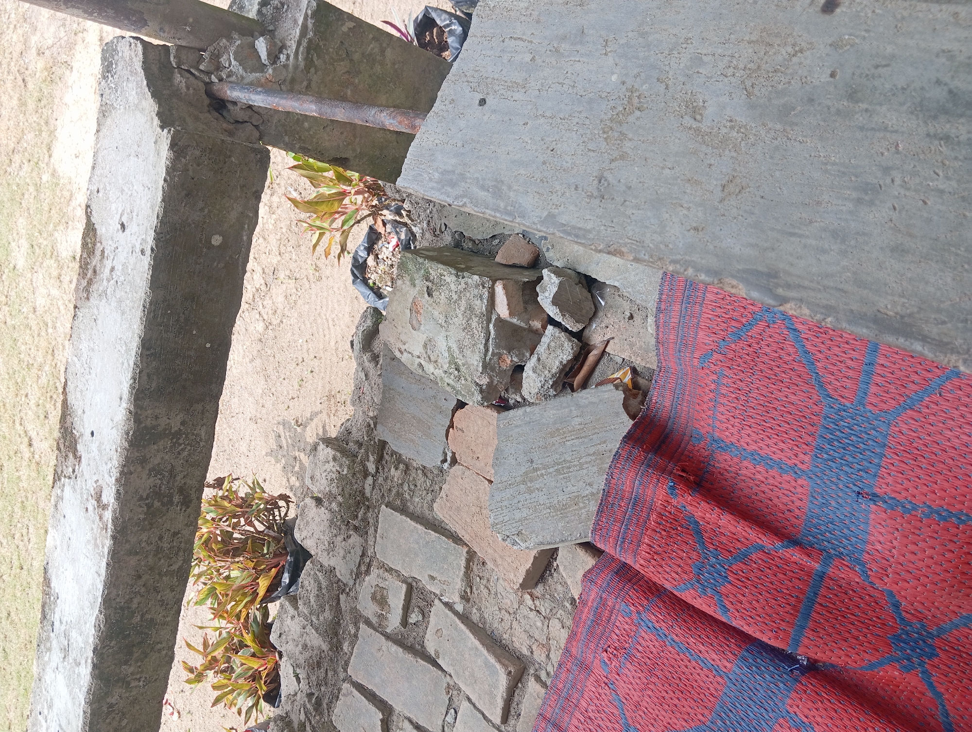 The image depicts an outdoor scene with concrete structures, plants in pots, and a red and blue woven mat. In the background, a grassy area is visible. The overall impression is of a construction or maintenance site, with debris and unfinished elements. The location is identified as Rejosari, Indonesia.