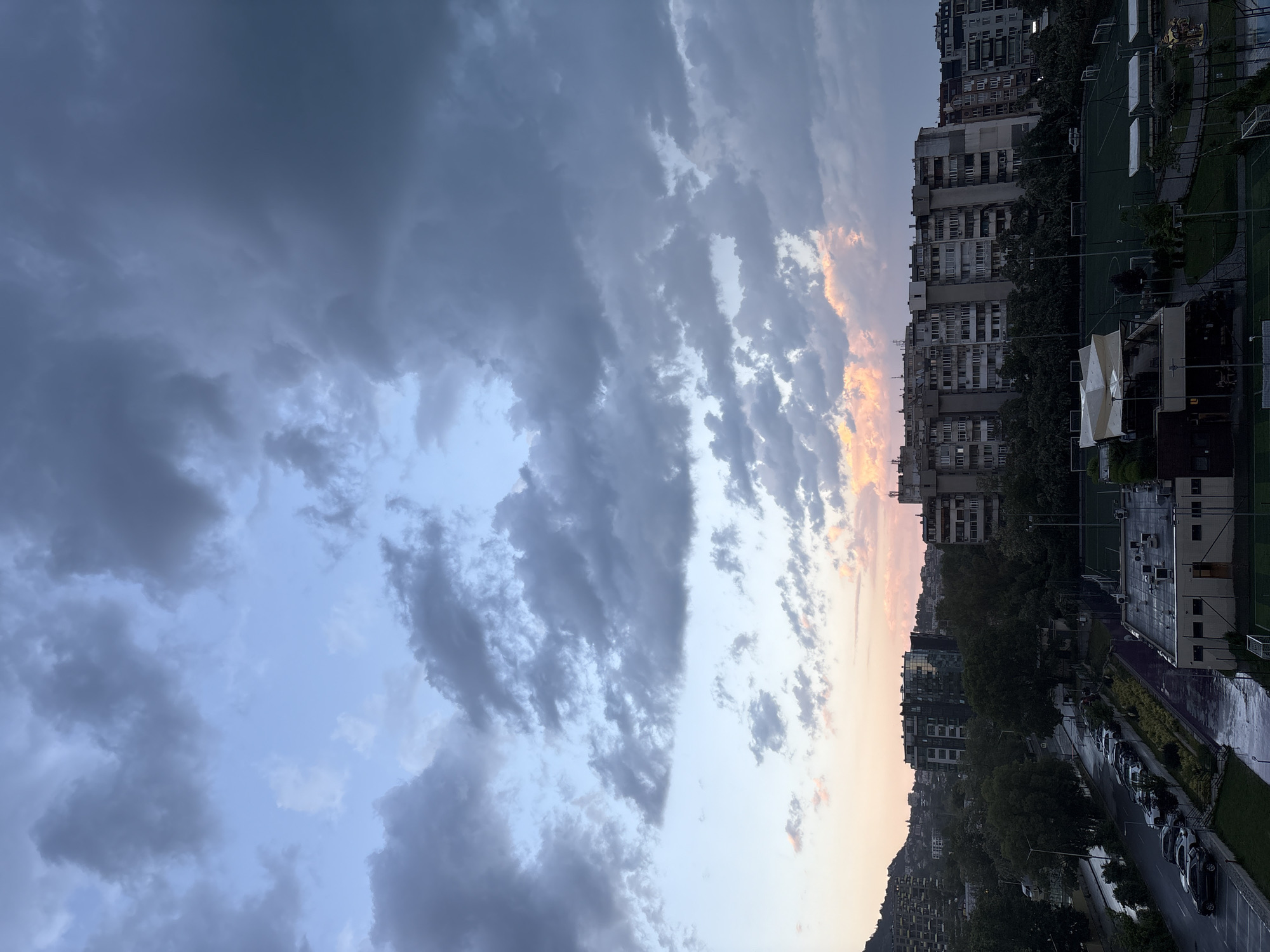 The image shows a skyline at dusk or dawn in an urban environment. Tall, multi-story residential buildings line the right side of the frame, some with visible windows and balconies. Adjacent to the buildings is a green sports field with artificial turf and white markings, suggesting it's used for soccer or a similar sport. Below the buildings, a street runs along the lower right corner, with parked cars visible. The sky dominates the image with layers of clouds, ranging in color from dark gray to light blue with some orange hues near the horizon, indicating either sunrise or sunset. The buildings and the field appear to be wet, possibly from recent rain. The location is in the city of Unknown, Unknown.