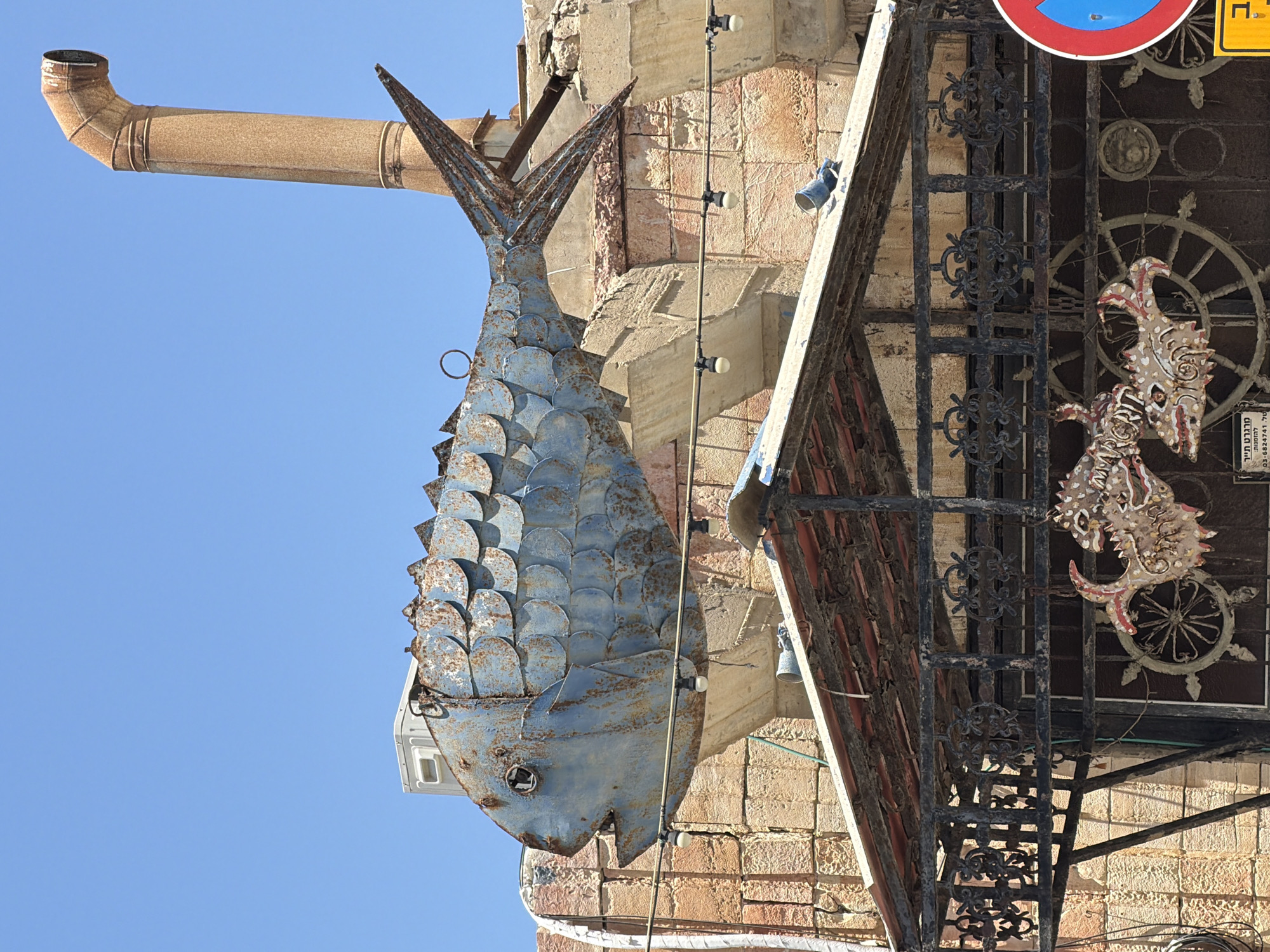 The image shows a corroded metal fish sculpture hanging outside a building in Tel-Aviv, Israel. The fish, made of blue-painted metal with individually crafted scales, appears weathered with rust and paint chipping. It is suspended from the building facade, near a ventilation pipe.
The building itself has a masonry exterior constructed with stone and brick. A black iron gate is visible next to the fish, adorned with decorative scrollwork. Behind the gate, another sign featuring a nautical theme, including a wheel and a mythical-looking creature is present. Various wires and light fixtures run along the building's facade. The sky is a clear blue.