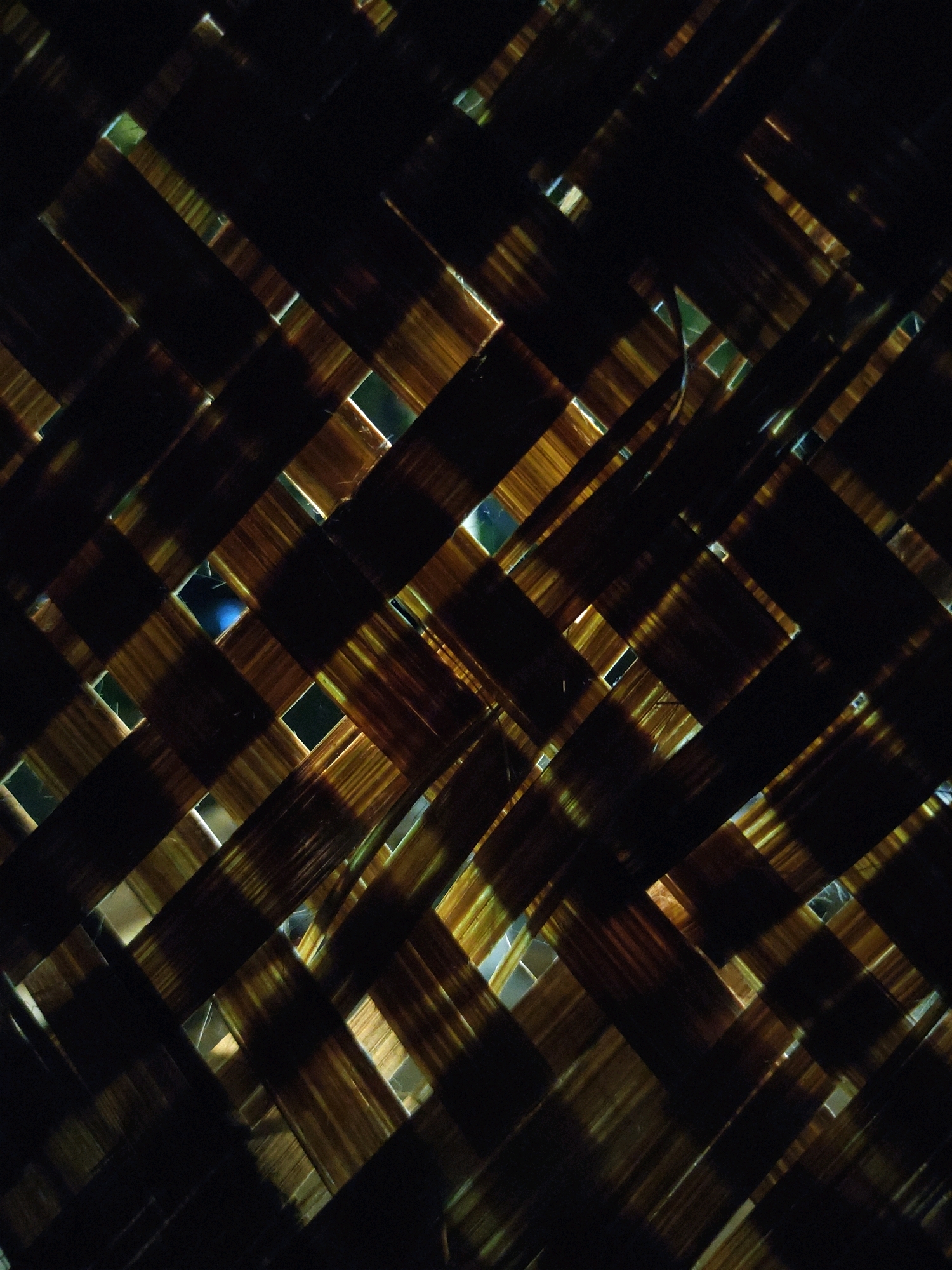 The image shows a close-up, abstract view of a woven mat, likely made from bamboo or palm leaves. The mat is dark brown and golden in color, with light filtering through the woven gaps, creating a pattern of bright rectangles and strips against the darker woven material. The lighting creates strong contrasts and highlights the texture of the material. There are no people, animals, or distinct objects visible. The image is a detail shot and does not provide information about its location in the city of Tiudan, Indonesia.