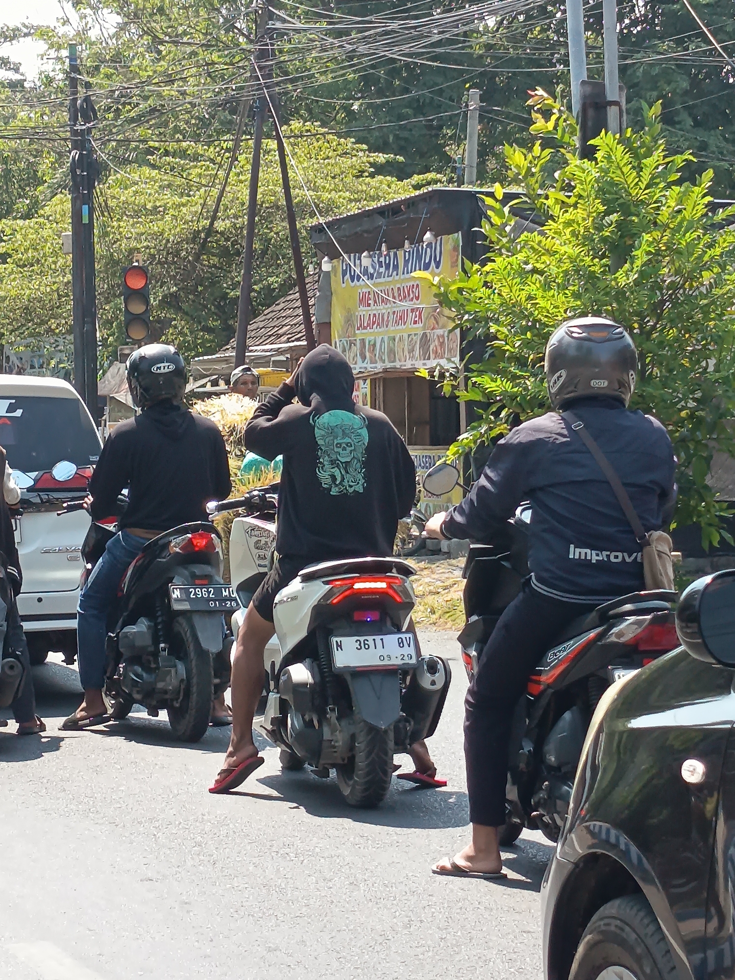 This image depicts a street scene in Kandangjati Kulon, Indonesia, on a sunny day. In the foreground, three individuals on motorcycles are stopped at a traffic light. The traffic light is red. The middle motorcycle has a license plate that reads "N 2962 MU 01-26". The motorcycle on the right has a license plate that reads "N 3611 QV 03-29". The person on the left motorcycle is wearing a black helmet and a black hooded jacket. The person in the middle is wearing a black hooded jacket with a skull graphic on the back and red flip-flops. The person on the right is wearing a dark blue jacket with "Improve" printed on the sleeve and black pants, also wearing a helmet. A white car is visible to the left. In the background, a building with a yellow sign advertising "SERA RINDO" and food items like "MIE AYAM & BAKSO" is visible. Trees and overhead electrical wires are abundant in the environment.