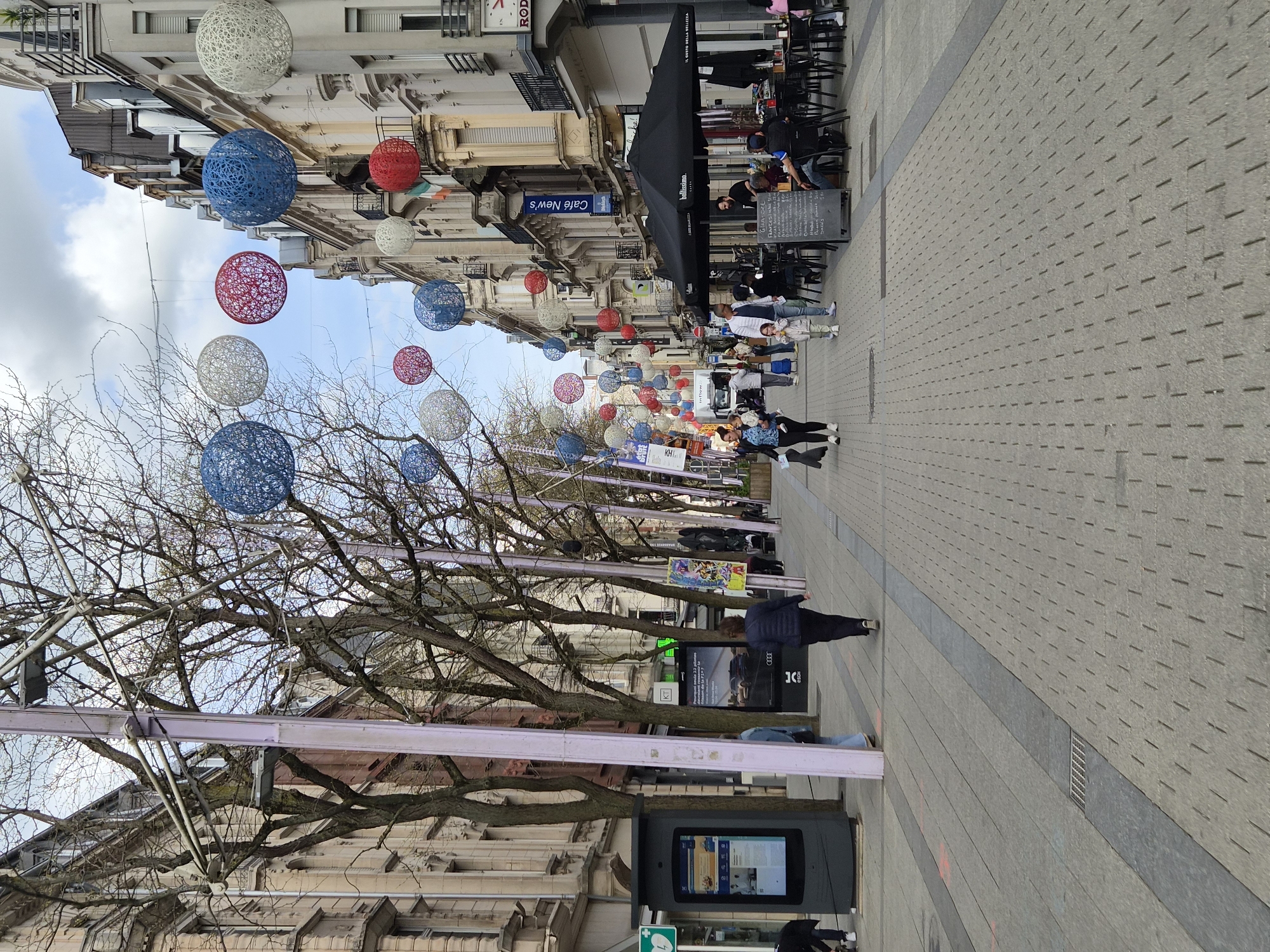 The image depicts a lively pedestrian street in Esch-sur-Alzette, Luxembourg, seen during the daytime under a partly cloudy sky. The street is lined with multi-story buildings housing various shops and cafes, showcasing European architectural styles.
Numerous large, decorative spherical lanterns, made of red, white, and blue interwoven strands, are suspended overhead, creating a festive ambiance. These colorful ornaments hang above the bare branches of trees that line one side of the street.
People are seen walking along the paved street, either individually or in small groups. On the right, an outdoor cafe is active, with patrons seated at tables under a large black awning that reads "Bellissimo." A menu board near the cafe entrance displays items like "GRAZZE," "FRANGO," "STEAK," and "SALADS." Visible signs for businesses include "Café New's," "LE CORPS DE LA BEAUTE," and a partially obscured "ROD."
Further down the street, an advertising stand features an "AUDI" logo, and a digital information kiosk displays various screens. The ground is paved with a light grey material, patterned with small rectangles and featuring a darker grey strip running down the center of the walkway. The overall scene suggests a casual, everyday atmosphere in a bustling commercial district.