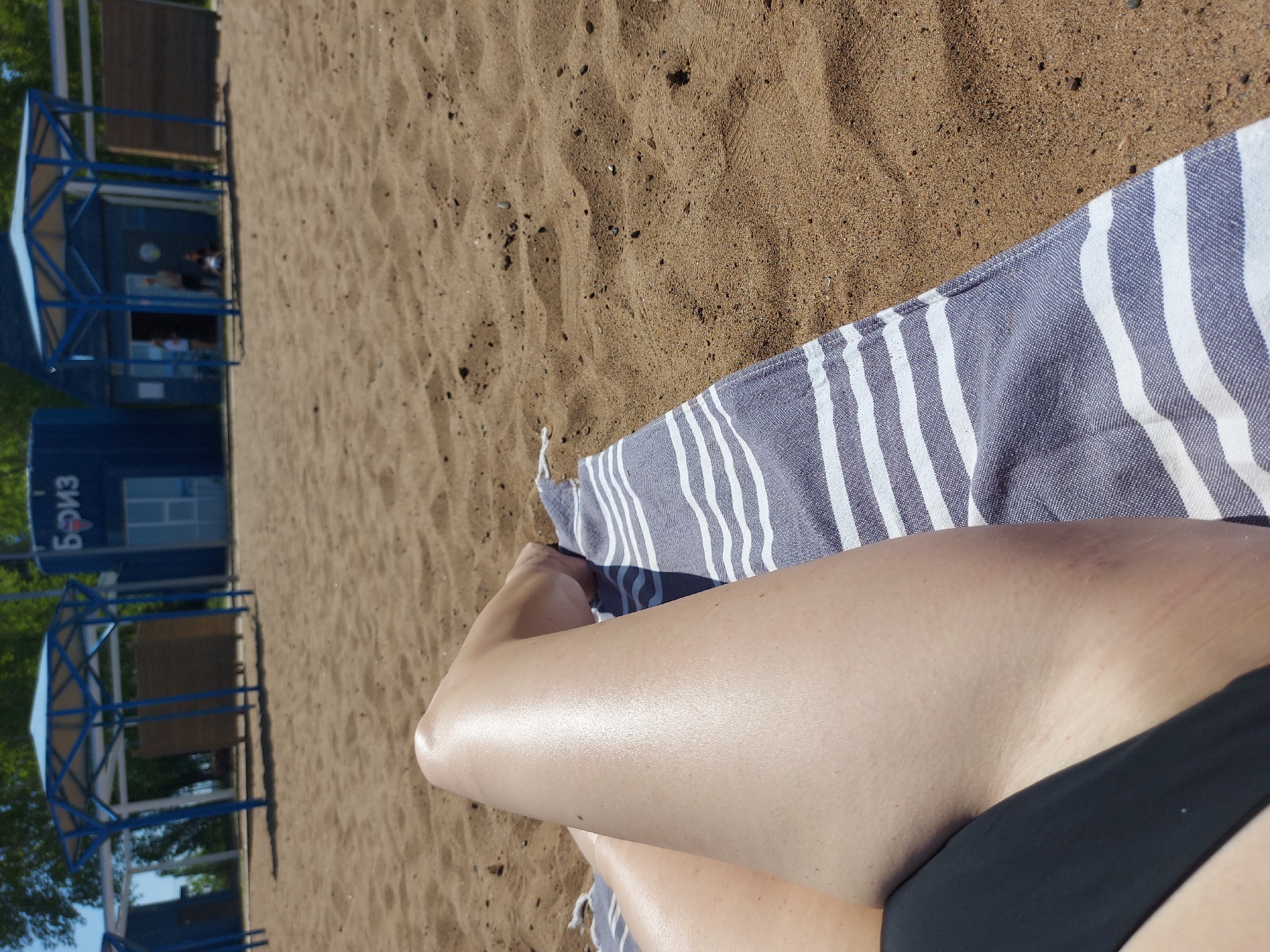 The image shows a person sunbathing on a beach in Chaikovsky, Russia. The beach is sandy and there are beach shelters with blue frames and roofs visible in the background. The person is lying on a blue and white striped towel. They are wearing black swimwear. The person's legs are visible in the foreground. The "Бриз" sign is seen on the building.