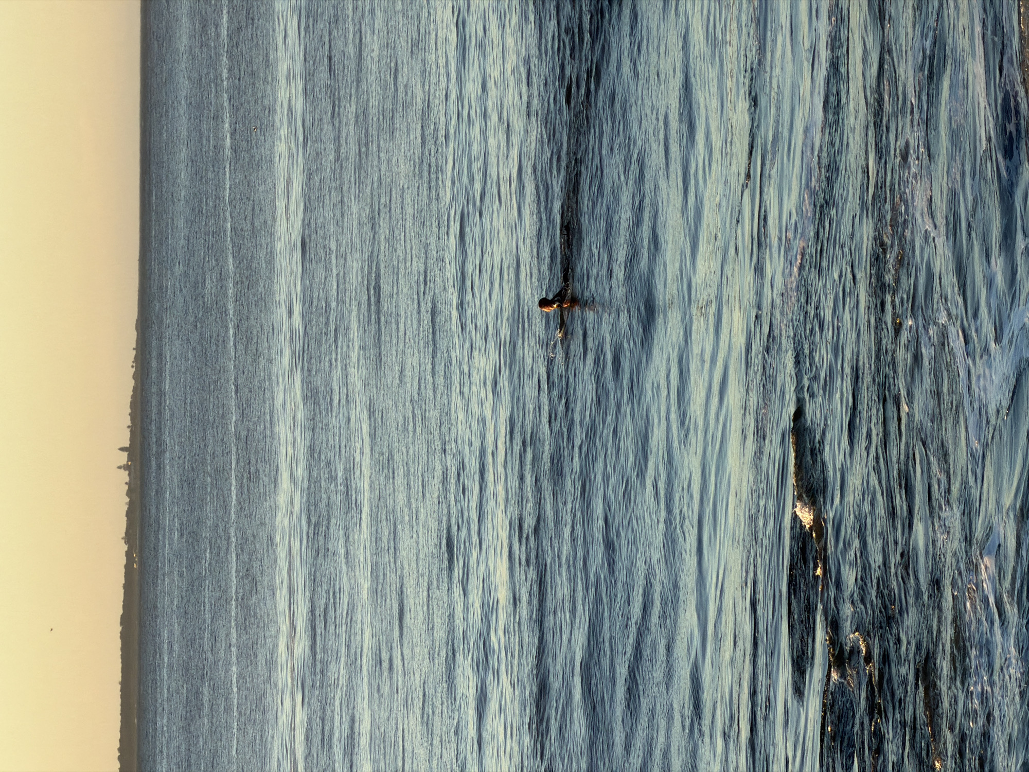 A lone swimmer, silhouetted against the calm ocean waters, is positioned in the mid-ground of the frame. The swimmer's head and shoulders are visible above the surface, and their body appears to be in motion, potentially swimming or treading water. The ocean is vast and stretches to the horizon, where a faint outline of land, possibly a city or coastline, is discernible against a soft, golden sky. The water exhibits gentle ripples, reflecting the warm hues of the setting sun. The scene evokes a sense of solitude and tranquility, with the individual swimmer emphasizing the expansive nature of the ocean. While the specific location in Cape Town, South Africa, cannot be definitively determined from the image alone, the overall seascape is consistent with the coastal environments found in and around the city, such as the Atlantic Seaboard or False Bay.