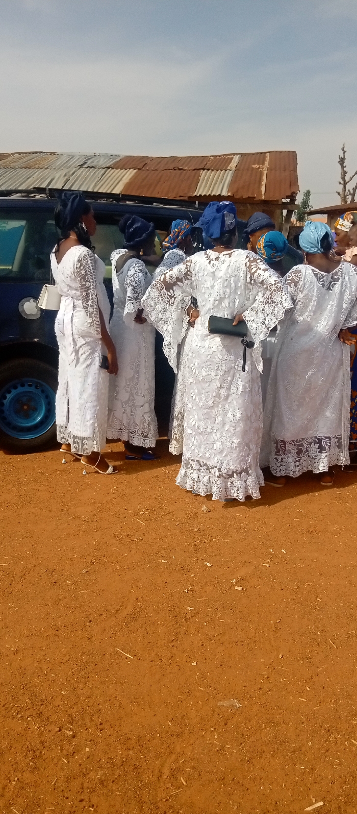 The image depicts an outdoor gathering in Kasuwan Magani, Nigeria, under a clear, pale blue sky. Approximately eight adult women are present, standing on reddish-brown dirt ground littered with small debris. All women are dressed in white, lace-patterned long-sleeve gowns, and many wear blue headwraps of varying shades. They are positioned adjacent to a dark-colored vehicle, likely an SUV or minivan, which displays an "Apple" logo decal on its rear side panel above a visible blue-rimmed wheel. In the background, a weathered structure with a rusty, corrugated metal roof is visible, along with sparse dry vegetation. Essential objects include a white shoulder bag held by a woman on the far left, a dark-colored clutch bag held by a woman facing away in the center, and mobile phones observed in the hands of at least two individuals. The women appear to be standing and engaging in social interaction.
