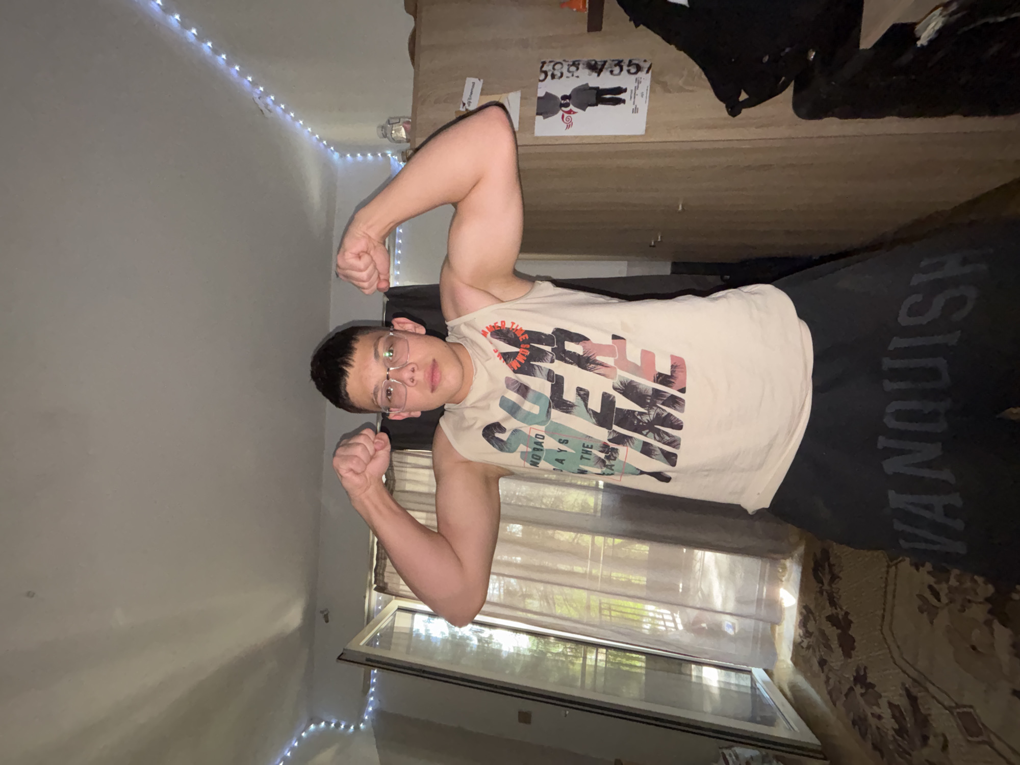 The image displays a young male posing indoors, flexing his biceps and looking directly at the camera. He wears clear-rimmed glasses and a light-colored sleeveless t-shirt adorned with a graphic featuring palm trees and text fragments like "NO BAD DAYS" and "SAVE THE WAVE." He is also wearing dark shorts that have the word "VANQUISH" printed on the left leg.
The setting appears to be a room, possibly a bedroom, illuminated by natural light from a large window or balcony door behind the subject, which reveals hints of trees or greenery outside through sheer curtains. To the person's right, a wooden furniture piece, likely a wardrobe or bed frame, is visible. A poster with the numbers "589 735" and "589 Y35" and a drawing of a person is attached to this furniture. Along the top-left wall, a string of blueish-white LED fairy lights provides ambient lighting. The floor is covered by a patterned rug in shades of brown. The lighting indicates it is daytime.