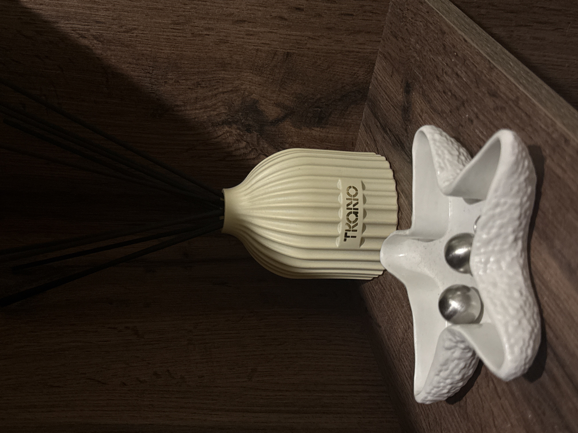 The image captures an indoor scene in Chaikovsky, Russia, featuring two decorative objects placed on a dark brown wooden surface.
On the left, a light beige or off-white aroma diffuser stands vertically, adorned with a ribbed, textured surface. Several dark, slender diffuser reeds protrude from its top, fanning out towards the left side of the frame. The brand name "TKONO" is embossed in a darker, contrasting shade on the side of the diffuser.
To the right of the diffuser, a white, star-shaped or abstract sculptural object is positioned. This object also has a textured, slightly bumpy surface, reminiscent of a coral or natural formation. Nested within two depressions of this white object are two spherical, shiny silver beads, reflecting the limited ambient light.
The wooden surface beneath the objects has a prominent grain pattern and appears to be a shelf or table. The lighting in the scene is dim, casting a shadow on the upper left portion of the wooden surface and creating a generally subdued atmosphere. The objects are well-defined, suggesting a focused light source from the upper right, while much of the background remains in shadow. No specific activity is depicted; the items appear to be static decorations.