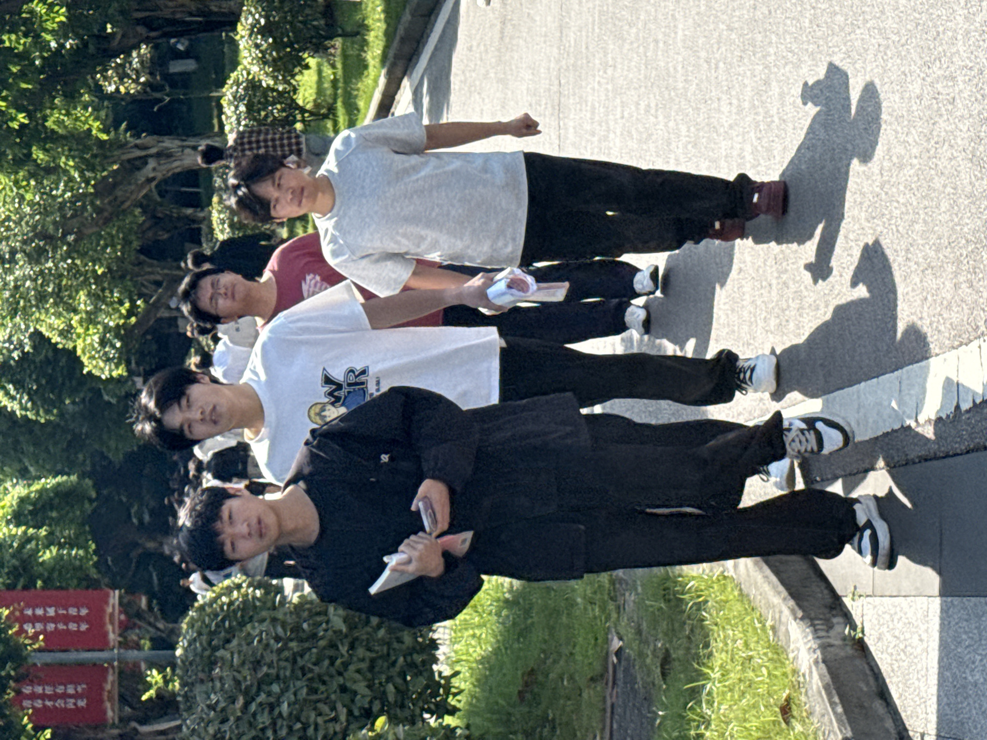 Four young men are captured in a daytime outdoor scene, walking or standing on a paved path lined with lush greenery and trees. The image, set in Jiangjin, China, shows them in casual attire under clear, sunny skies, indicated by the prominent, elongated shadows cast on the light-colored pavement.
From left to right, facing the viewer:
The first man wears a black long-sleeved shirt or jacket, black pants, and black and white sneakers. He is holding a white object, possibly a smartphone or small book, and a small red rectangular item in his hands.
The second man wears a white short-sleeved t-shirt with a graphic displaying "MR" and cartoon-like figures, black pants, and white sneakers.
The third man wears a red short-sleeved t-shirt, black pants, and white sneakers. He holds a white and red packet, possibly tissues or a small snack.
The fourth man wears a light grey short-sleeved t-shirt, black pants, and maroon/brown shoes. His right arm is bent, with his hand clenched in a fist.
The background features additional out-of-focus individuals and dense foliage. On the far left, a red rectangular sign with vertical Chinese characters is partially visible, reinforcing the location in China. The overall mood appears relaxed, with neutral to slightly smiling expressions on the men's faces.