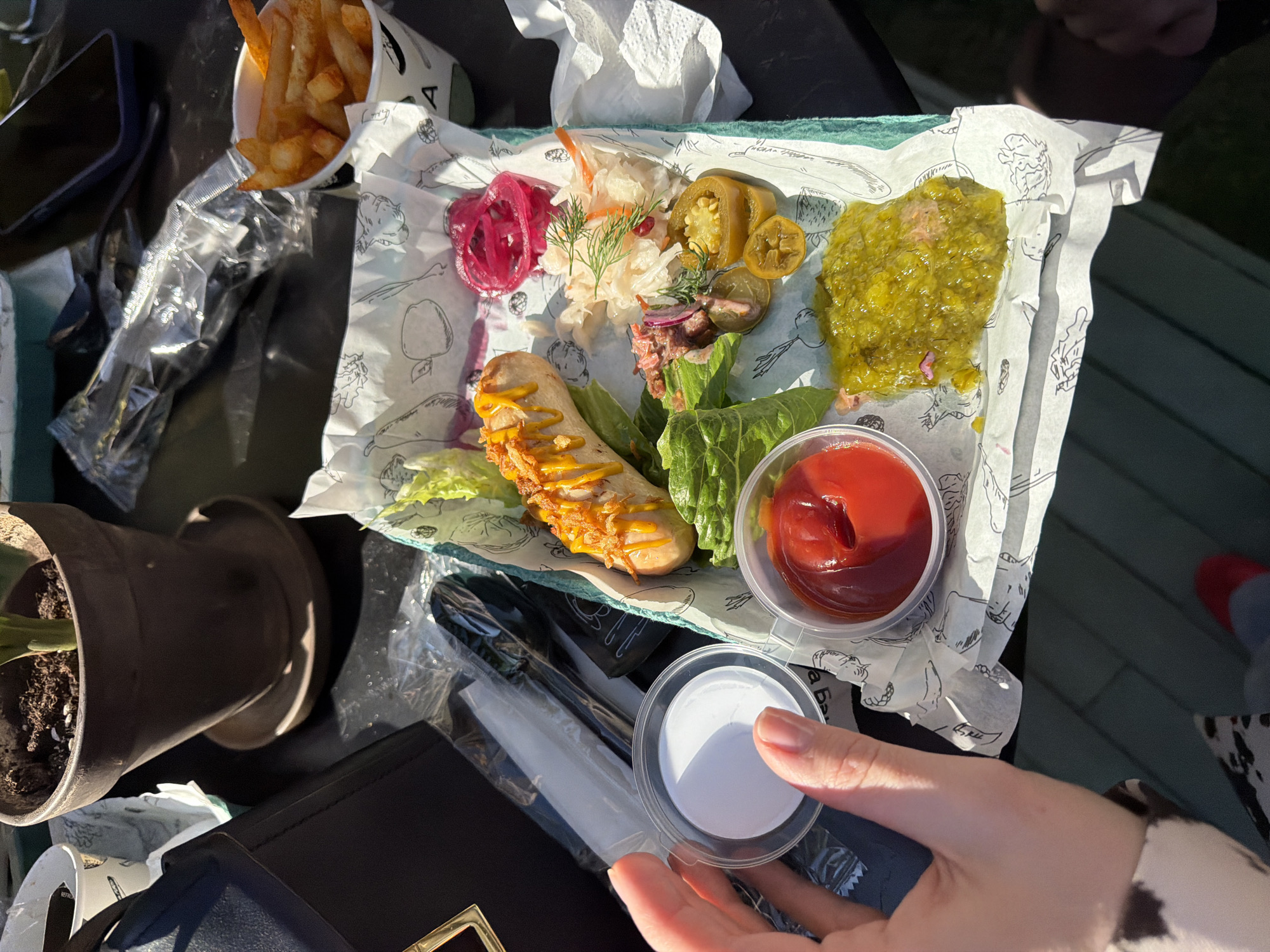 The image shows a tray of food, including a sausage with mustard, pickled vegetables, a condiment of some sort, and a side of fries. The tray sits on an outdoor table, along with a potted plant, a phone, and a black purse. A hand is holding a small container, possibly containing another condiment. The location and details about the city of Moscow, Russia, are not discernible from the image.
