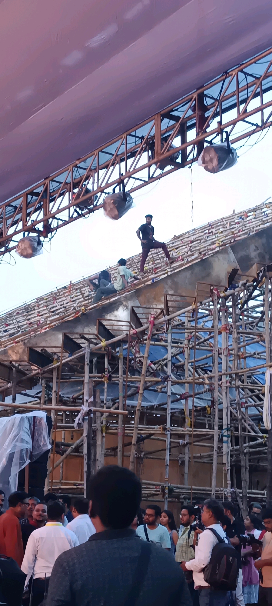 In Khardaha, India, a group of men are working on the construction of a large, temporary structure, likely for a festival or event. The structure appears to be built primarily from bamboo scaffolding and wooden planks, with a large fabric canopy overhead. Several spotlights are suspended from the scaffolding. Three men are visible actively working on the elevated part of the structure, which has a sloped roof covered in colorful decorative elements. Below, a crowd of people are gathered, some appearing to be onlookers or participants in the event. The scene suggests a dynamic environment of ongoing construction and anticipation.