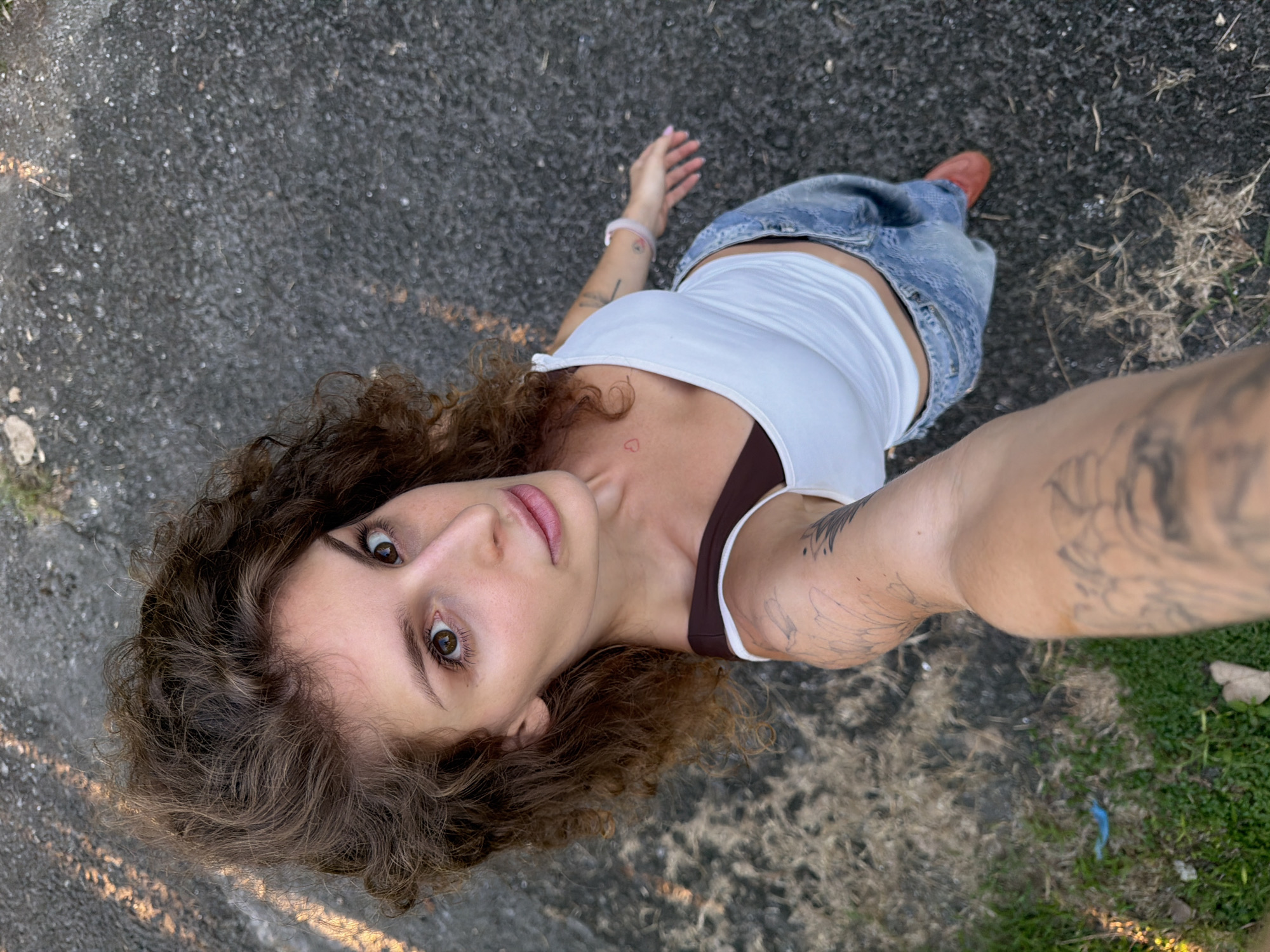 A woman is angled upwards in a self-portrait photograph. She has light skin, brown curly hair, and brown eyes. She is wearing a white tank top with brown straps, loose-fitting denim jeans, and one red shoe is visible. Her arm, closest to the camera, is outstretched with tattoo art visible. A heart tattoo is visible on her chest near her collarbone. The background appears to be asphalt and some grassy areas. The image location is indeterminate and cannot be identified as Kediri, Indonesia.