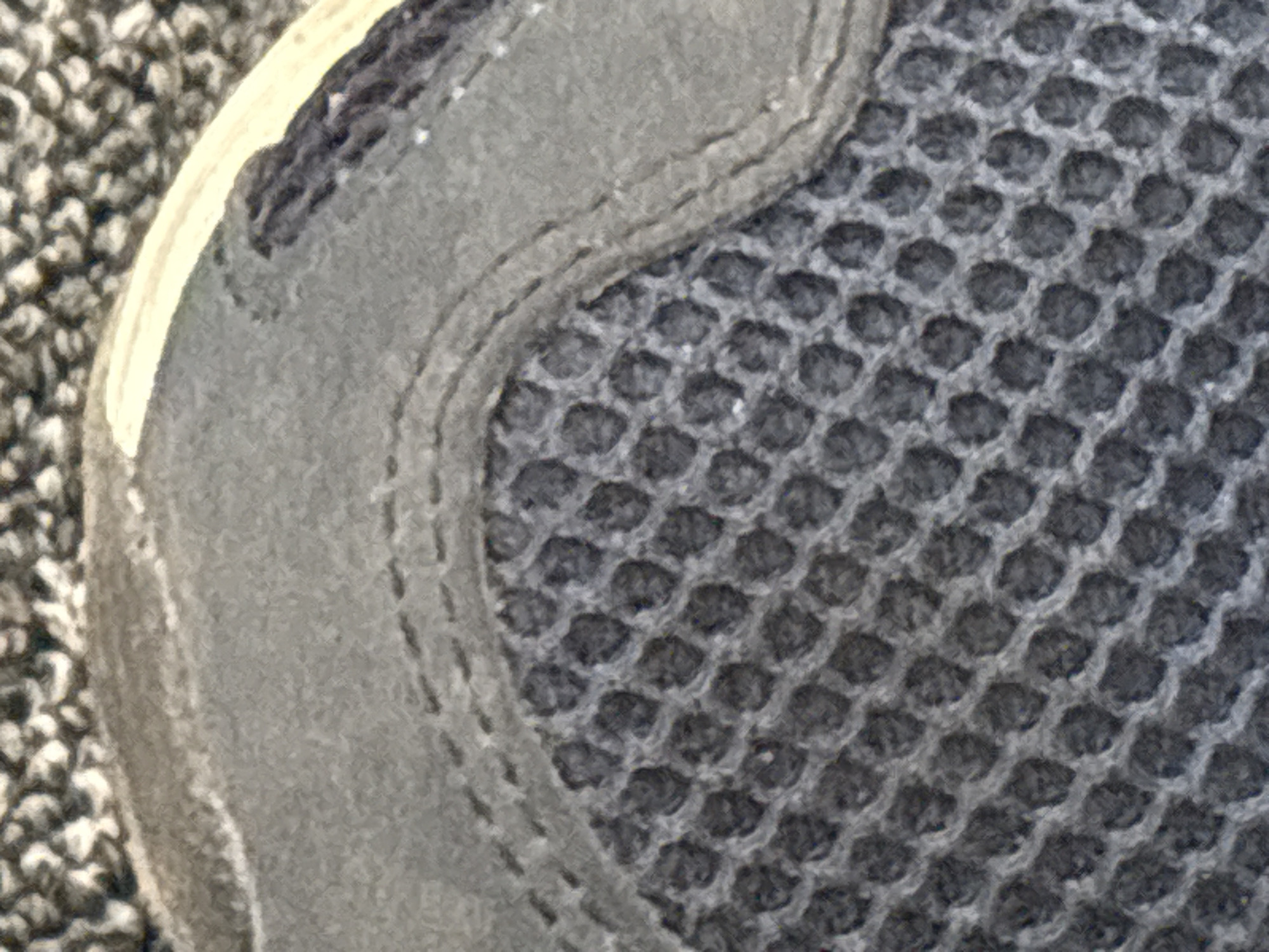 The image is a close-up, oblique, high-angle, full shot of a grey mesh athletic shoe. The shoe material is a dark grey woven mesh with a honeycomb pattern. A lighter grey suede-like material overlays the mesh, with visible stitching. A light tan strip runs along the top edge of the shoe. The shoe is resting on a textured surface that appears to be carpet or a similar material, with light and dark grey fibers. There are no people or other identifiable objects present. The scene is indoors, given the presence of carpet, but the specific location within a city is unknown.