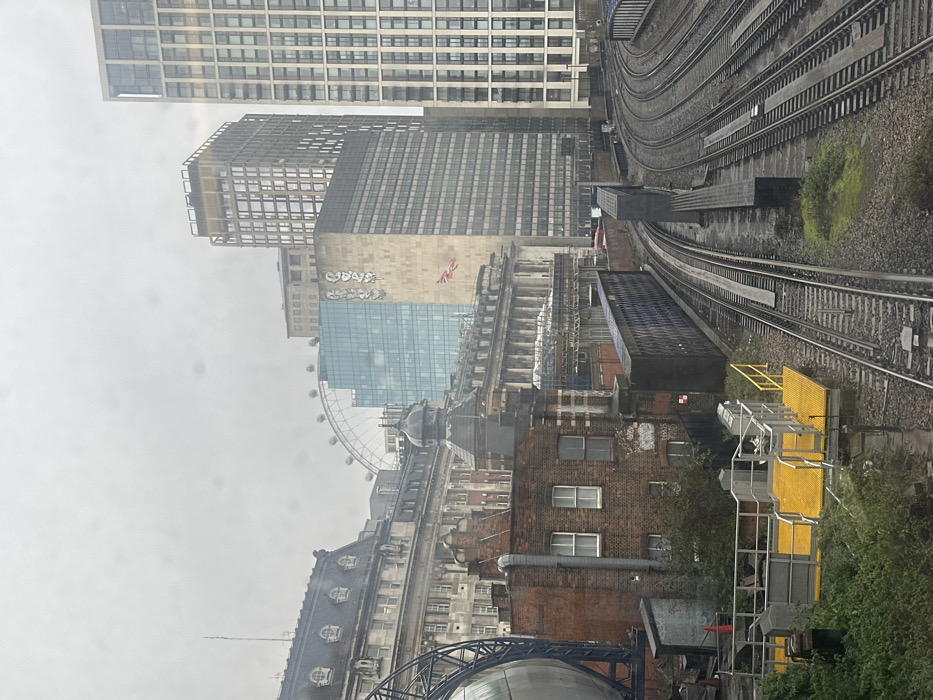 The image presents an elevated, panoramic view of railway tracks and urban architecture in the Waterloo area of London, United Kingdom, under an overcast sky.
In the foreground, numerous parallel railway tracks, appearing wet from recent precipitation, extend into the midground, set in gravel ballast. Green foliage grows sporadically alongside the tracks. On the far left, a bright yellow elevated platform with grey railings and a small grey control box is visible. Dark grey, rectangular metal structures, likely railway bridges or retaining walls, are positioned between and alongside the tracks.
The midground features a mix of historical and modern buildings. On the left, a large, spherical metallic structure within a dark framework is partially visible, indicative of the BFI IMAX cinema. Behind it, a classical-style, multi-story building with a mansard roof and ornate details stands prominently. Below this, an older, reddish-brown brick building with multiple rectangular windows and an external grey pipe is situated directly adjacent to the railway lines. To its right, another ornate stone building with a dark domed roof and classical columns is visible.
The background is dominated by the London skyline. The London Eye Ferris wheel is clearly visible on the left. Adjacent to it, a modern, light-blue glass-fronted building (London Television Centre) stands next to a taller, beige-colored corporate building (IBM South Bank) featuring a large white logo near its top and a Union Jack flag hanging from its facade. To the right, two prominent, tall, slender, modern residential towers with numerous windows rise into the sky, one showing signs of ongoing construction at its apex. No individuals or specific actions are depicted. The scene is static, captured on a damp, grey day.