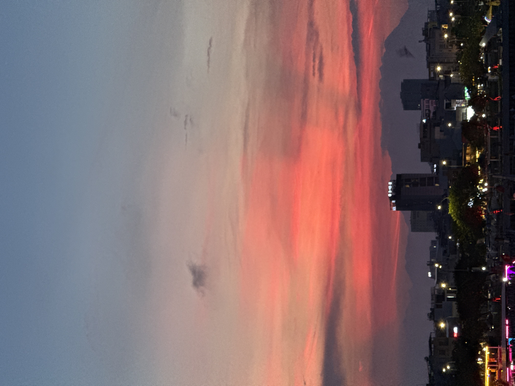 This image captures a vibrant sunset over the city of Da Nang, Vietnam. The sky is ablaze with fiery reds and oranges, transitioning to soft pinks and blues towards the top. Silhouetted against the colorful sky are distant mountains, softened by a light haze. In the foreground, the city skyline is visible, with buildings of varying heights and architectural styles. Streetlights are beginning to illuminate, casting a warm glow on the urban landscape. Some trees are also visible, their foliage dark against the twilight sky. The overall impression is one of a peaceful evening scene in a coastal city.