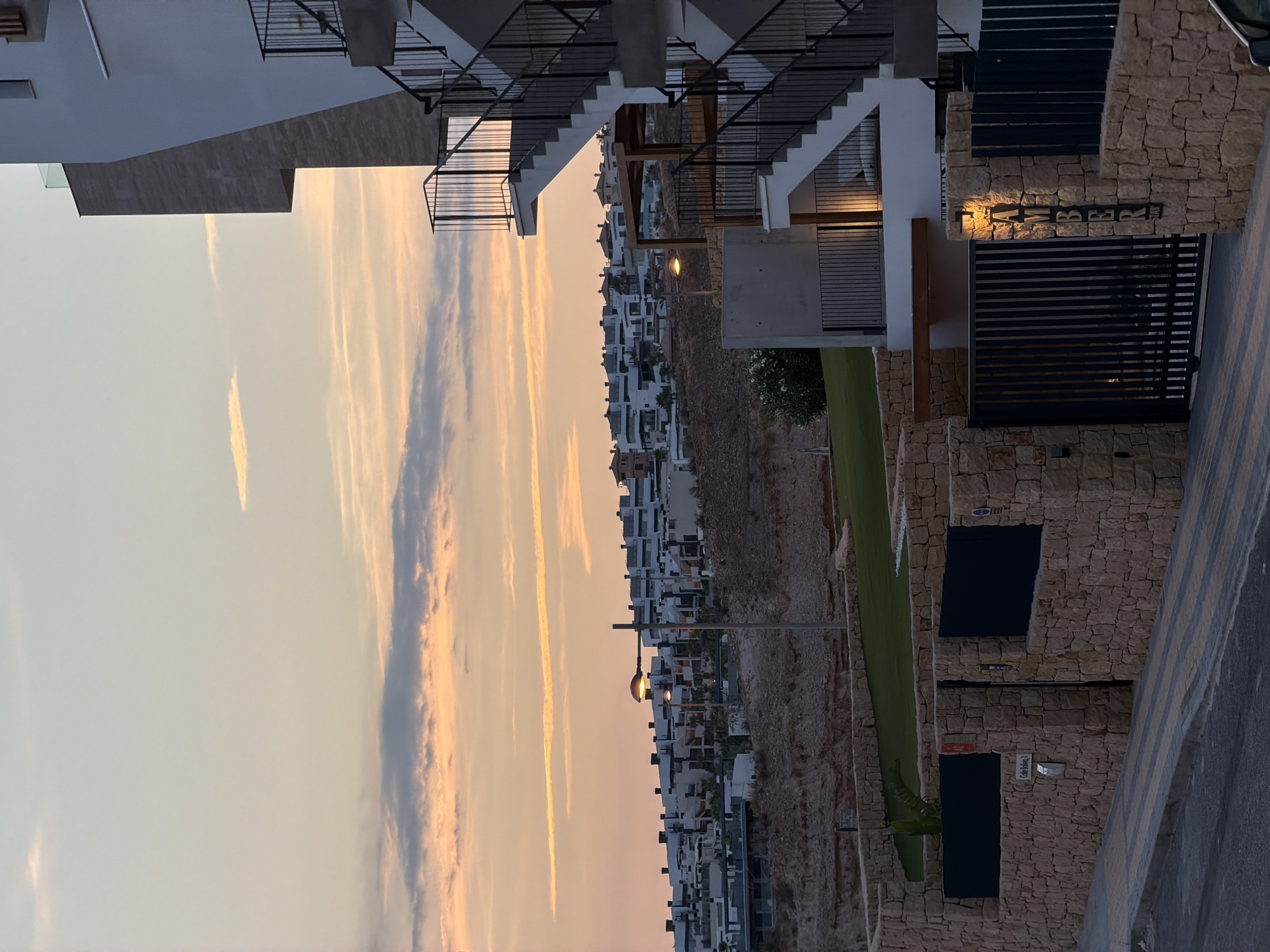 This media file, captured in Finestrat, Spain, depicts a modern residential complex built into a hillside. The architecture features white buildings with dark accents, many with balconies and staircases. The complex is set against a backdrop of a rocky incline and a sprawling urban landscape dotted with similar white structures stretching towards the horizon.
The scene is illuminated by the warm glow of sunset, casting long shadows and painting the sky with hues of orange, pink, and pale blue. Wispy clouds streak across the sky, adding to the serene atmosphere. A streetlamp is visible, its warm light also contributing to the evening ambiance. The overall setting suggests a tranquil evening in a Mediterranean coastal town.
The image is oriented vertically, with the sky occupying the left half of the frame and the buildings and hillside on the right. The right side of the image shows a stone-clad wall with a dark metal gate and the word "AMBER" visible in raised lettering above the gate. Other small signs are also present on the stone walls, though the text is mostly illegible. The presence of greenery, including what appears to be a lawn and some bushes, adds a touch of life to the constructed environment. The overall mood is peaceful and picturesque, highlighting the blend of modern architecture with the natural landscape at dusk.