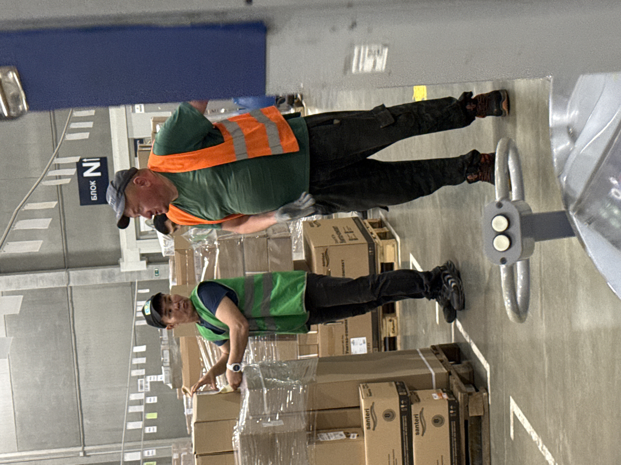 In Lupanovo, Russia, two male workers are inside a warehouse. The worker on the left, wearing a blue t-shirt, green vest, and black cap, is examining a stack of boxed goods wrapped in plastic. He has a watch on his left wrist. The worker on the right, wearing a green t-shirt, orange safety vest, and dark pants, is standing and looking to his left. He is wearing gloves and appears to be engaged in conversation. The environment is a large industrial space with concrete floors and high ceilings, with numerous stacks of boxes and pallets visible throughout the area. A blue pillar is present to the right of the frame.