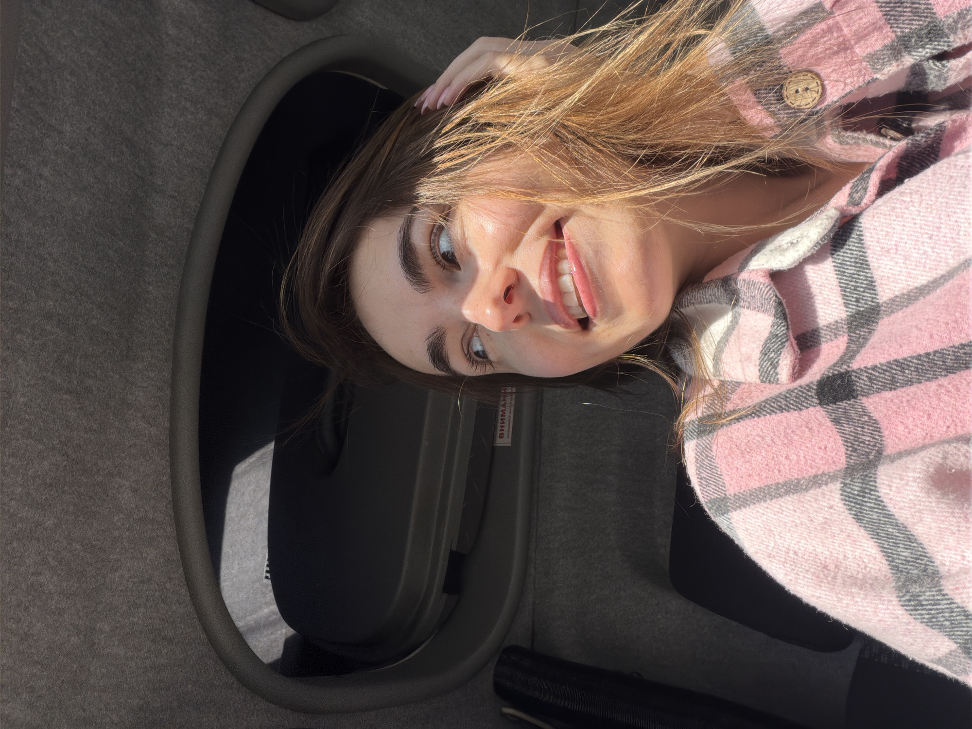 A woman is lying down in what appears to be the trunk of a car. She is fair-skinned with light brown hair and is wearing a pink and gray plaid flannel shirt. Her head is tilted toward the camera and she is smiling, with one hand resting on her hair. The trunk interior is gray and black, with a cylindrical black object visible in the lower portion of the frame. A label with Russian text is affixed to the side of the trunk. The location is not discernible from the image.