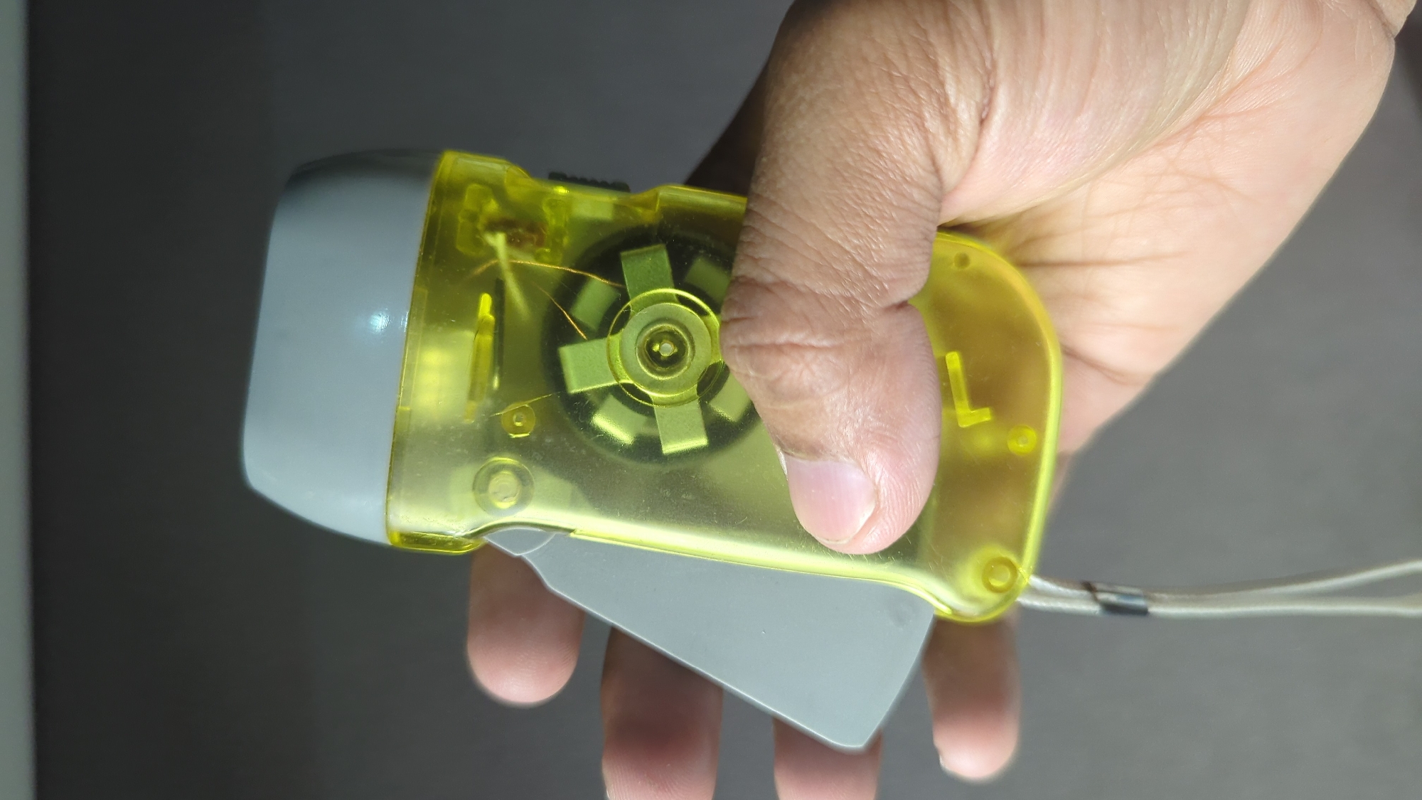 In the city of Penjor, Indonesia, a close-up shot focuses on a hand holding a yellow, transparent hand-crank flashlight. The flashlight has a grey cap at the top, and a glimpse of its internal workings, including a star-shaped rotor, is visible through the translucent casing. A light-colored lanyard is attached to the bottom of the flashlight. The background is a neutral, blurred grey, suggesting an indoor or dimly lit environment.