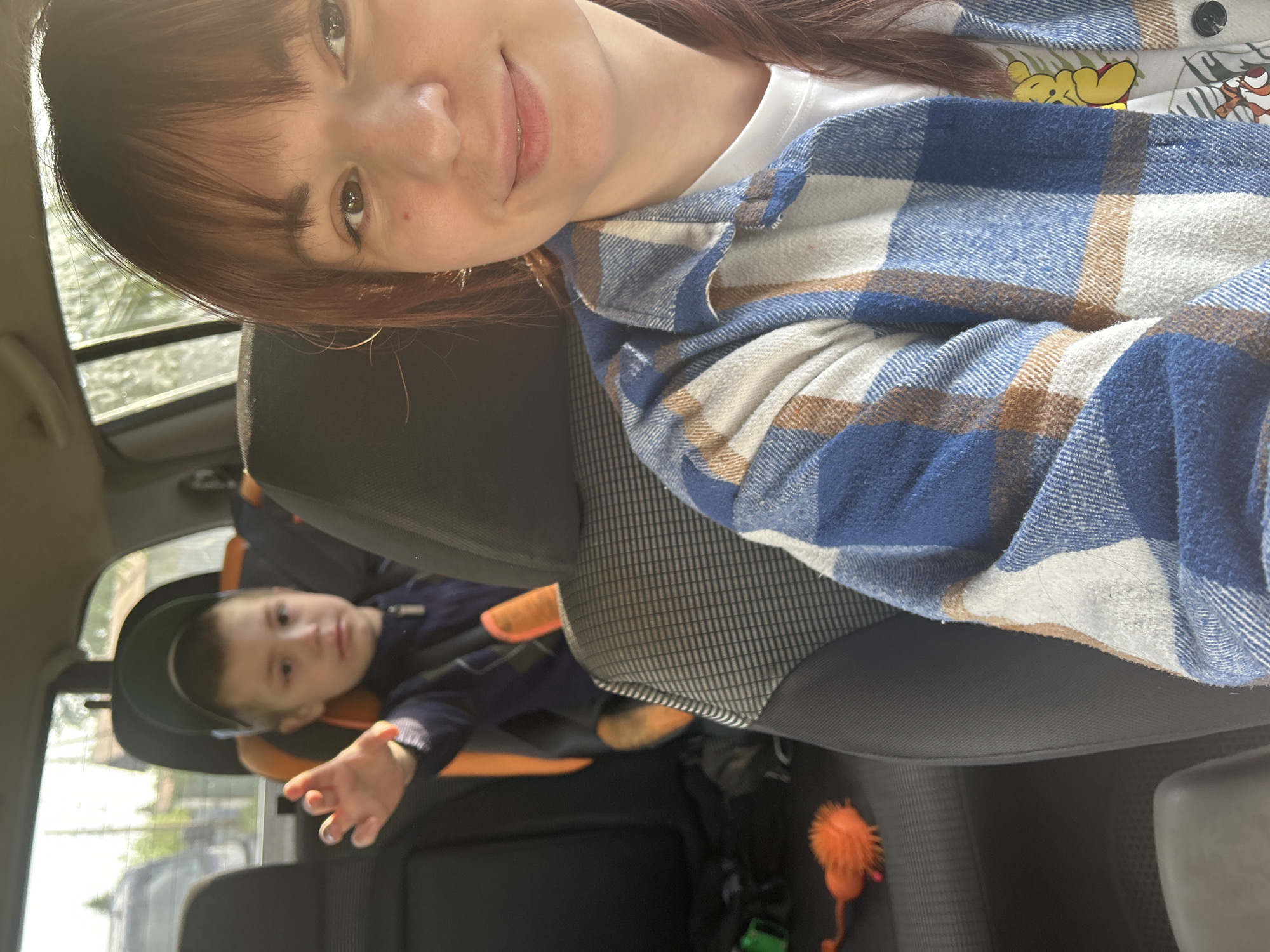 A woman with brown hair and a blue and white plaid shirt is taking a selfie inside a car. In the back seat, a young boy is sitting in a car seat. An orange fuzzy toy is on the car floor. The location cannot be determined to be in the city of Фоки, Russia based on this image.