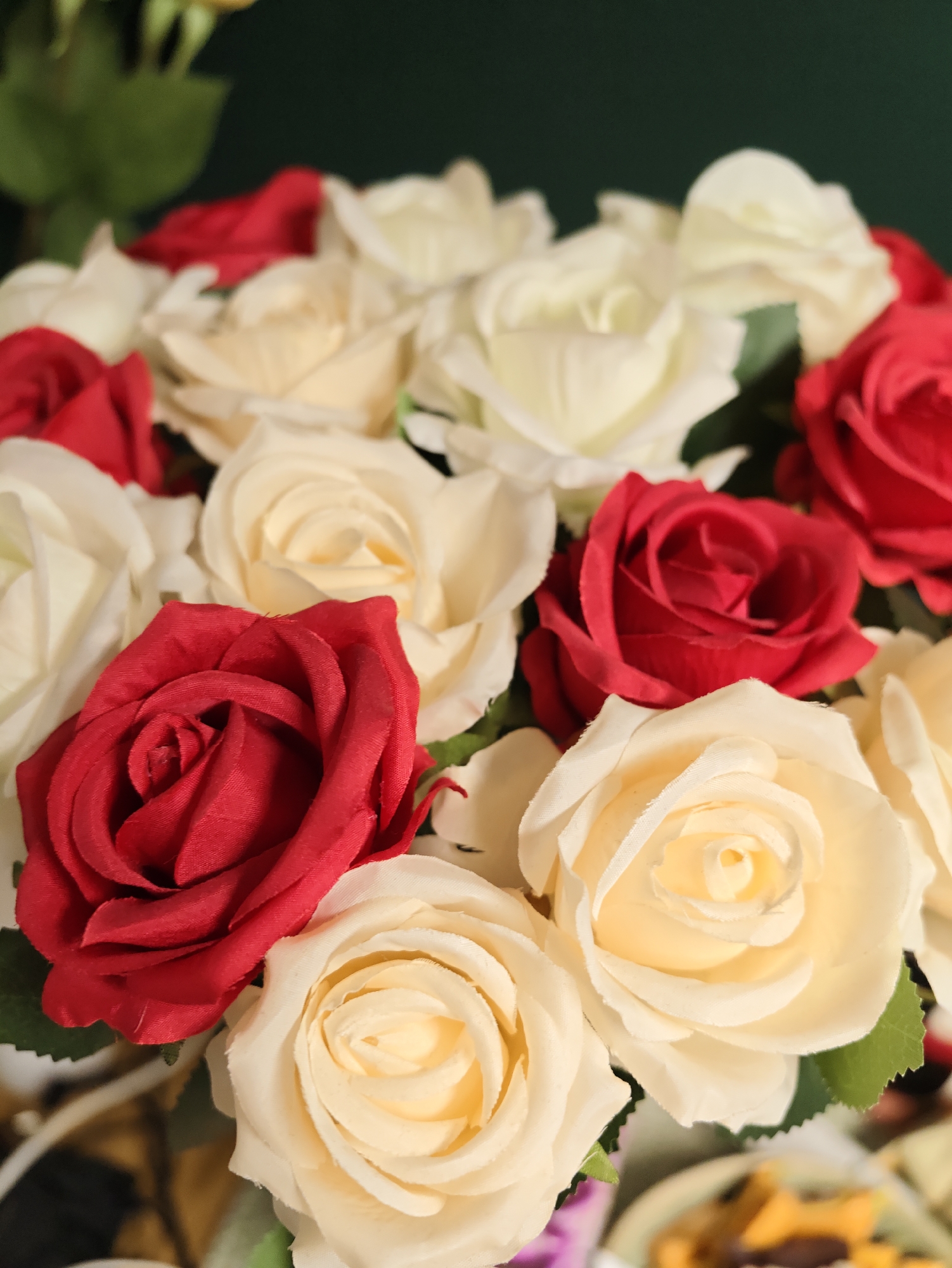 A bouquet of artificial roses, a mix of red and cream colored blooms, is arranged in a close-up shot. The roses appear to be made of fabric, with realistic petal textures. In the background, a dark green surface is visible, possibly a wall or foliage, adding contrast to the vibrant colors of the flowers. The lighting is soft, highlighting the delicate folds of the rose petals. The image does not provide any information about its location in Petah Tikva, Israel.