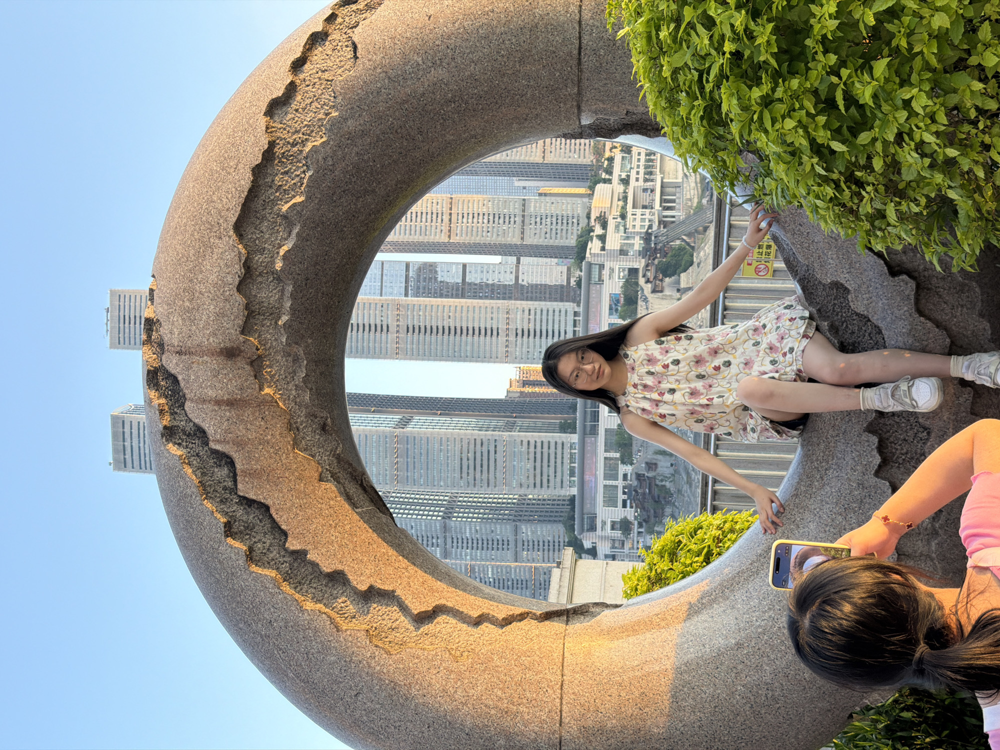 In Nan'an District, China, a young woman with glasses and long dark hair is seated on a large, rough-hewn stone sculpture that resembles a circular archway. She is wearing a floral print sleeveless dress and white sneakers. Her gaze is directed forward, and her arms are extended slightly. In the foreground, another person is taking a photo of her with a smartphone. This second person has dark hair pulled back in a ponytail and is wearing a pink shirt. The sculpture is situated outdoors, with lush green foliage visible to the right. Through the circular opening of the sculpture, a cityscape with numerous tall, modern buildings is visible under a clear blue sky. The sunlight casts a warm glow on the sculpture, highlighting its textured surface.