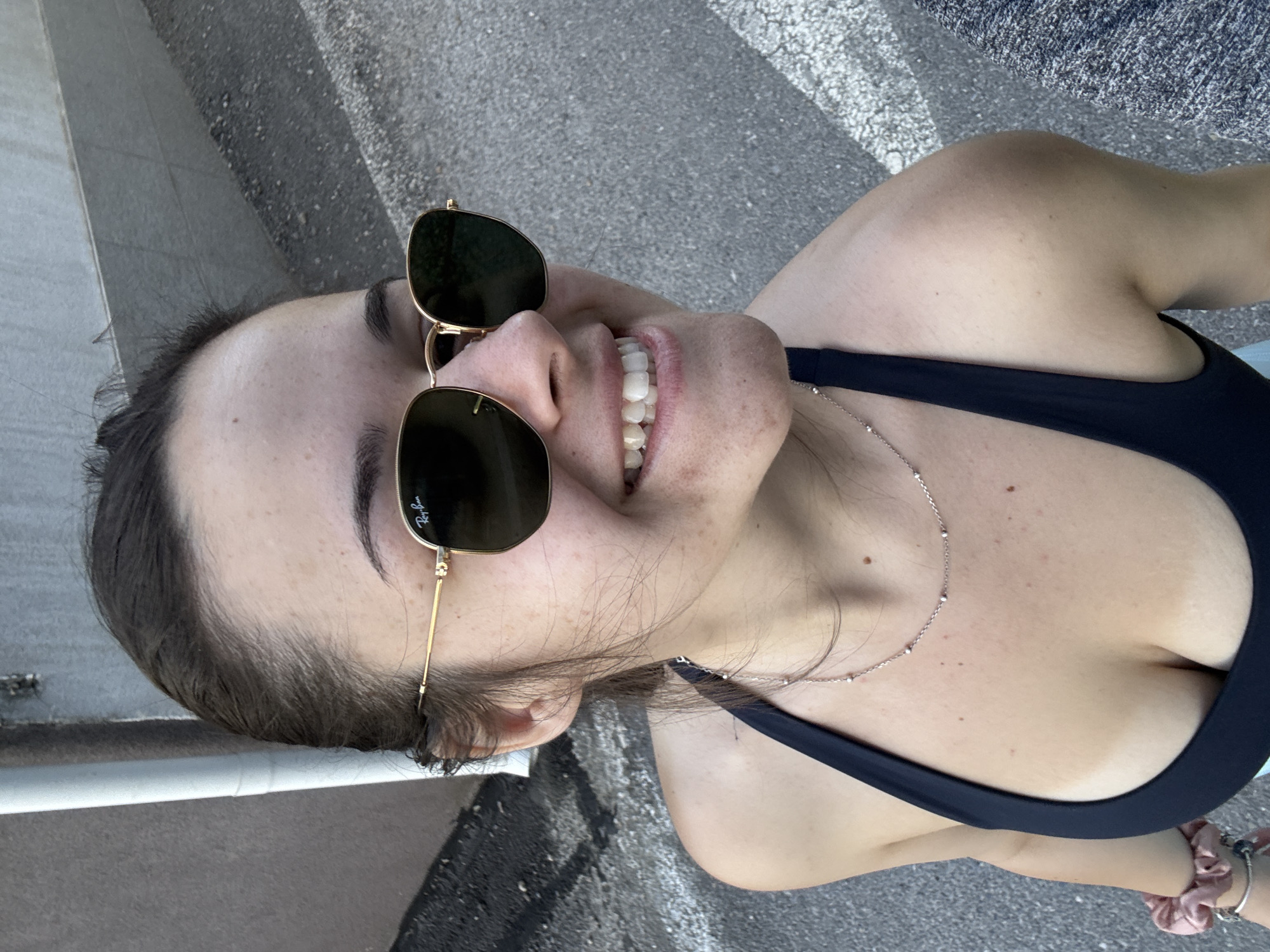 A woman with brown hair pulled back and wearing dark sunglasses is smiling and looking up at the camera. She is wearing a black sports bra and a silver necklace. Her arm is visible with pink scrunchies around her wrist. The background consists of an urban setting with concrete and a wall, likely in a city. It is difficult to confirm that the location is Sofia, Bulgaria, without additional context or clues within the image.