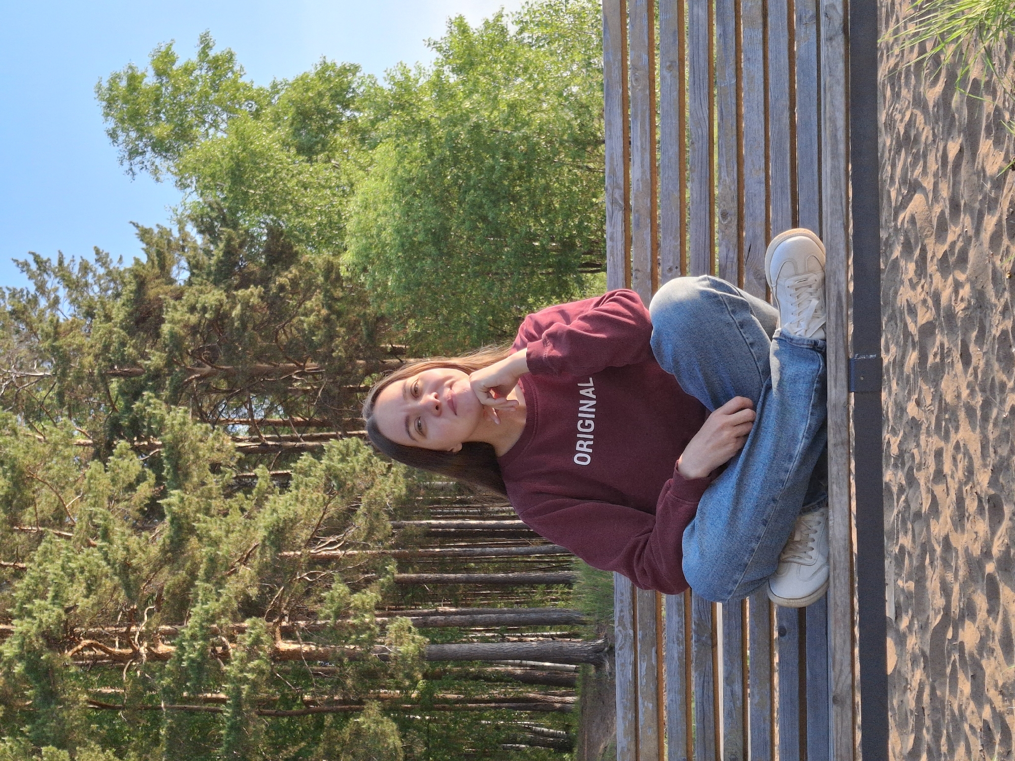 A medium shot shows a young woman sitting on a wooden bench with a backdrop of trees, possibly located in Chaikovsky, Russia. The woman is wearing jeans and a maroon sweater with the word "ORIGINAL" printed on it. She is leaning on her hand, gazing towards the camera. The environment includes a combination of coniferous and deciduous trees, and sand to the side of the bench. The sky is blue.